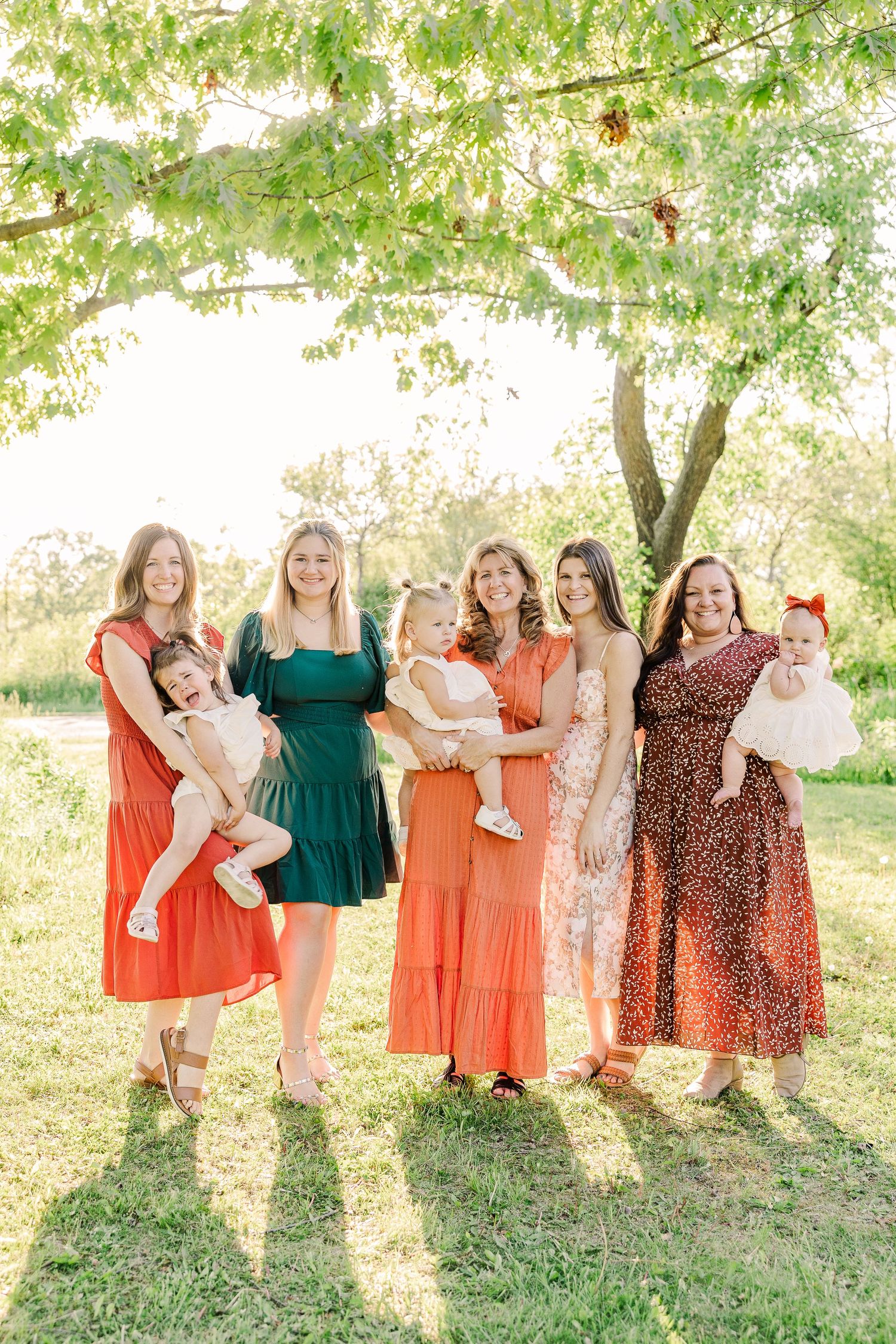 Group of friends in coordinating sundresses gather under a tree in warm sunlight during an outdoor photoshoot.