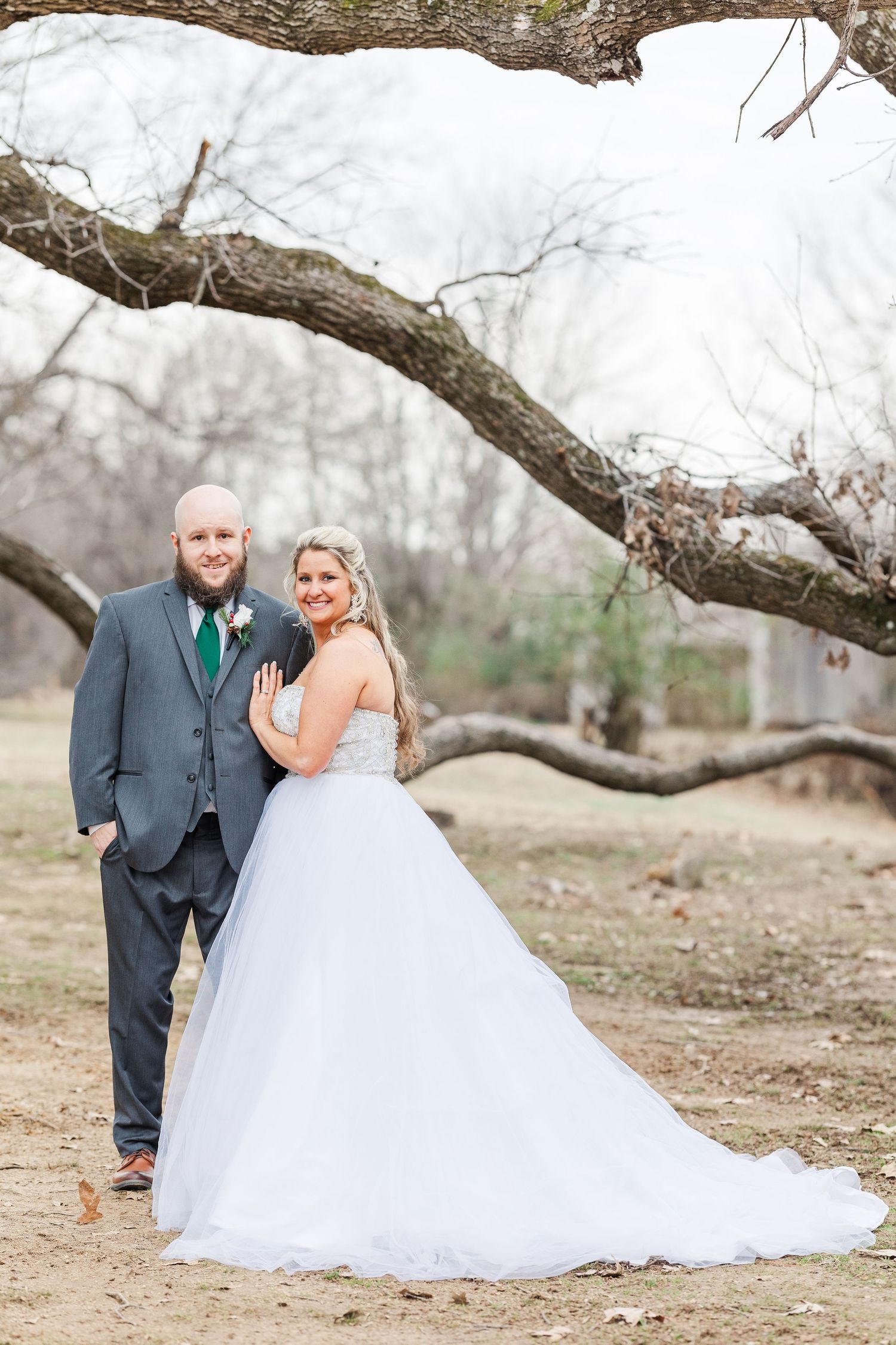 Wedding couple poses beneath a dramatic curved tree branch in a winter woodland setting.