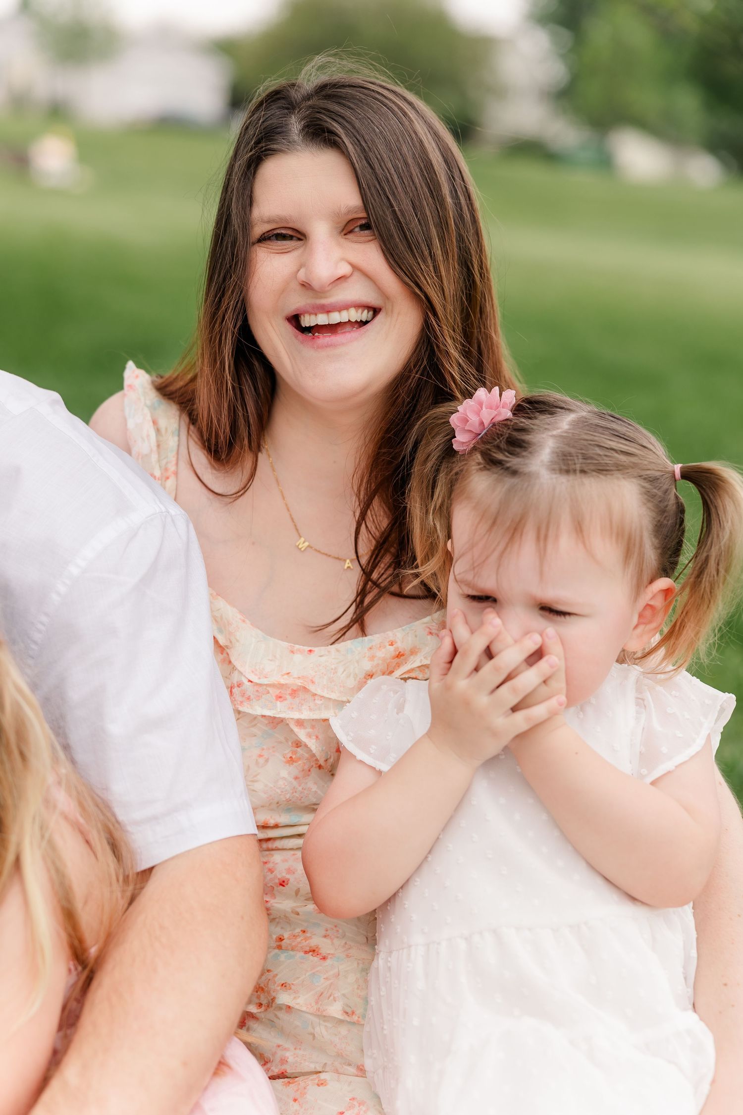 Laughing young brunette woman with smiling toddler girl in white dress during outdoor family photo session.