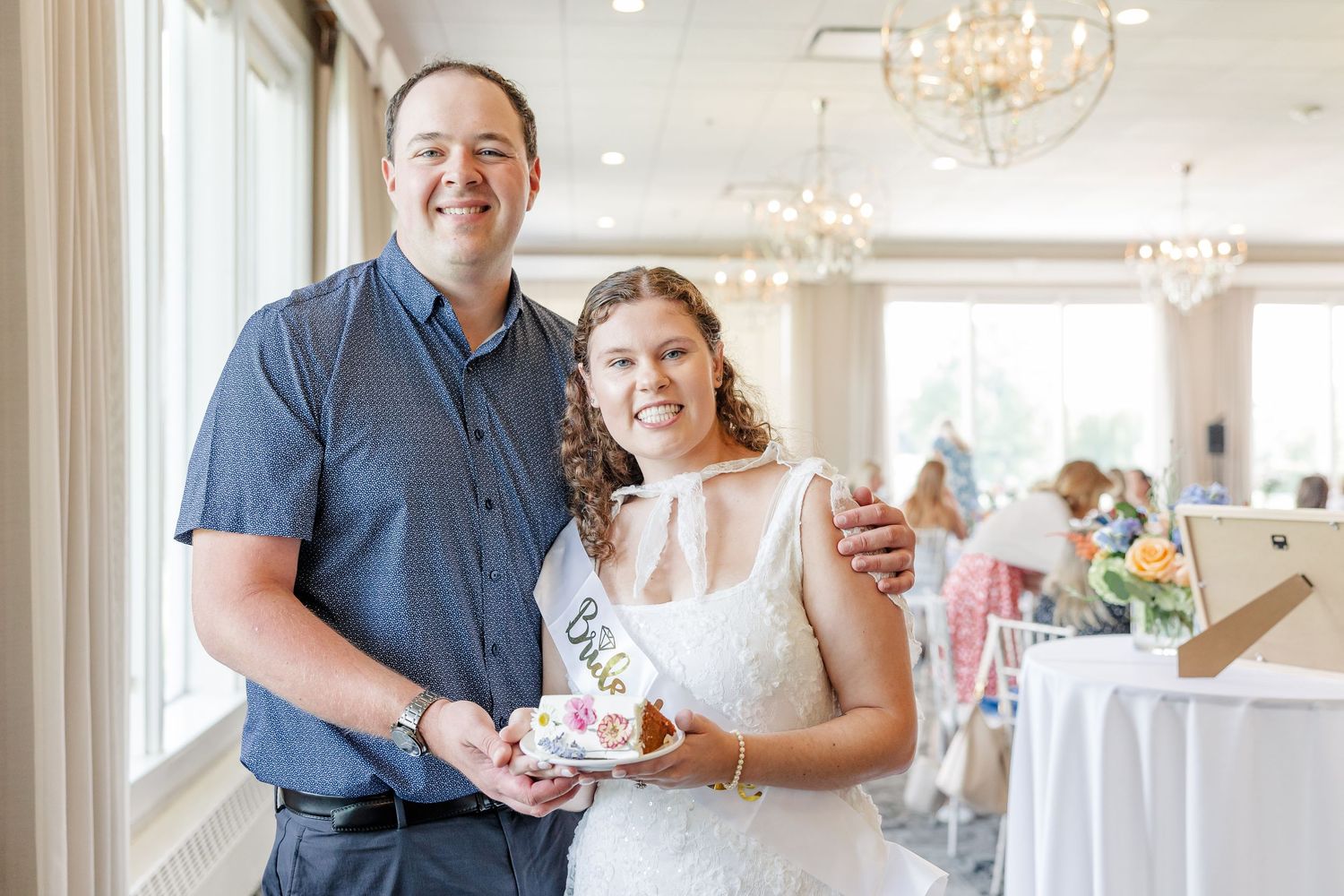 Smiling couple at a bridal shower, woman wearing a Bride sash holding a small gift indoors.