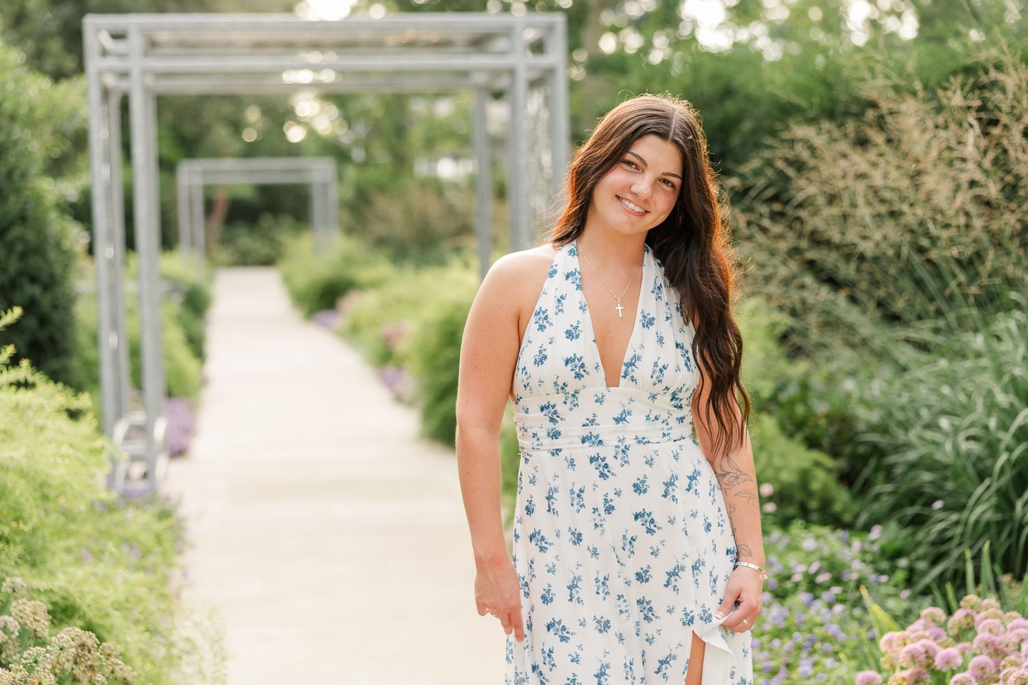 Young woman in white floral halter maxi dress poses in a lush garden pathway with green foliage.