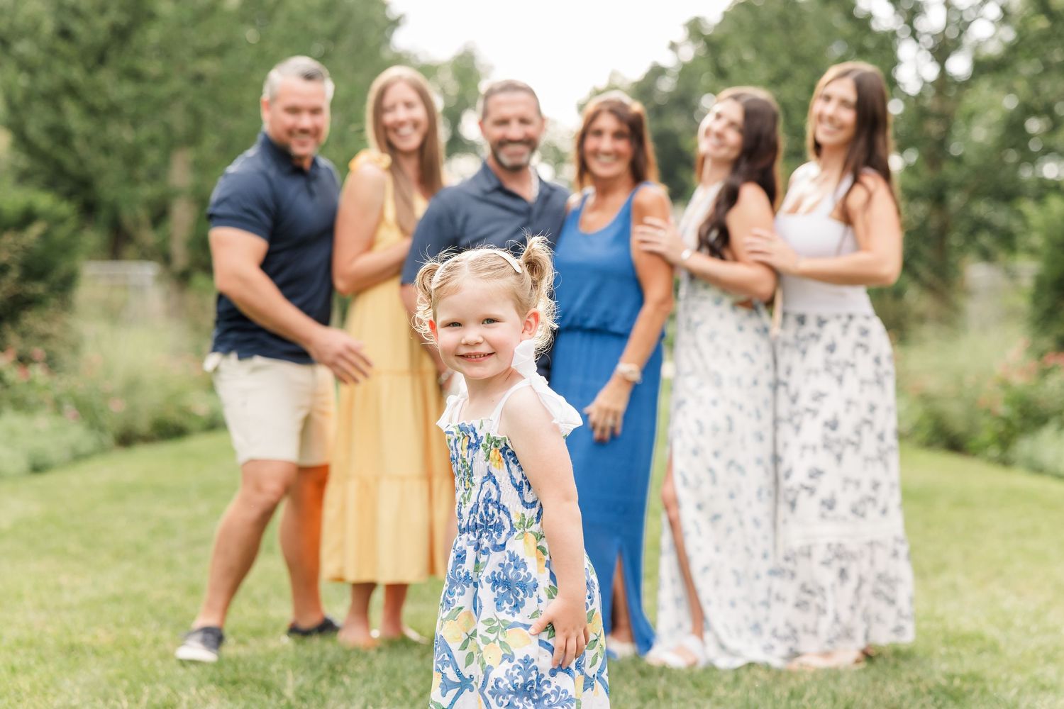 Young girl in blue floral dress smiles in foreground with family of five posing on green lawn behind her.