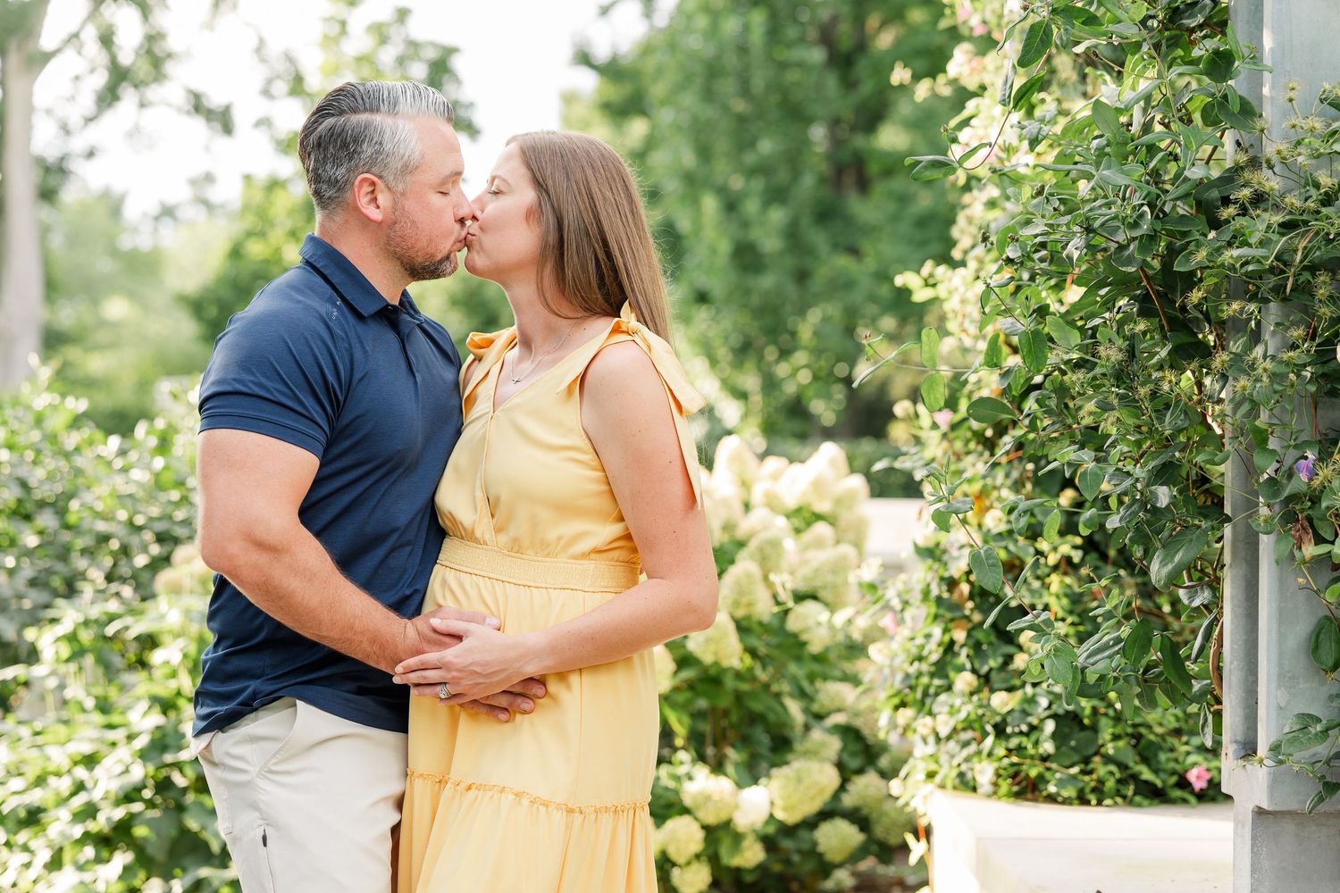 Couple sharing a tender kiss near white flowering vines, man in navy polo, woman in yellow dress.