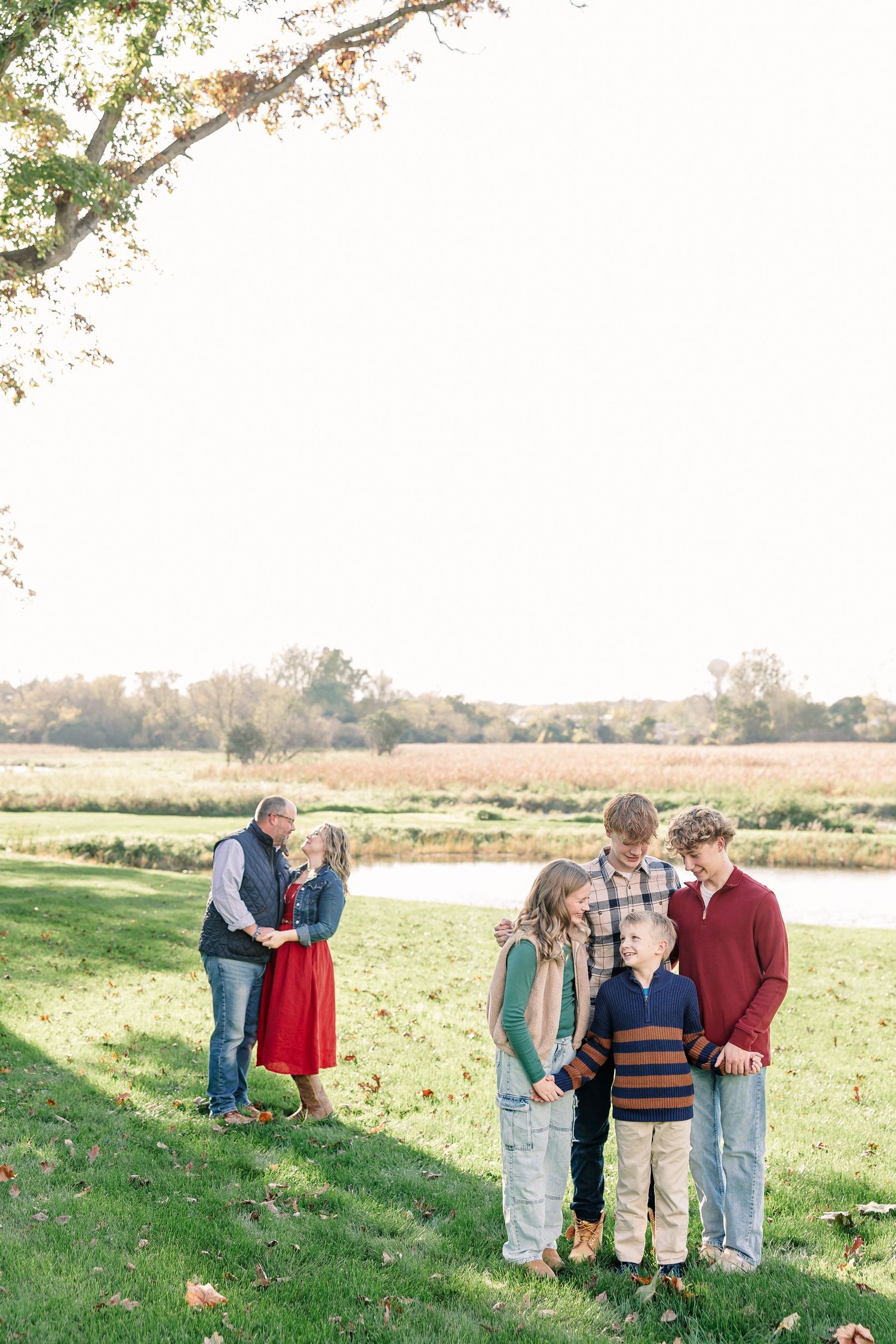 A family of six poses on a grassy lawn by a pond in autumn, with a couple embracing while four children stand together nearby.
