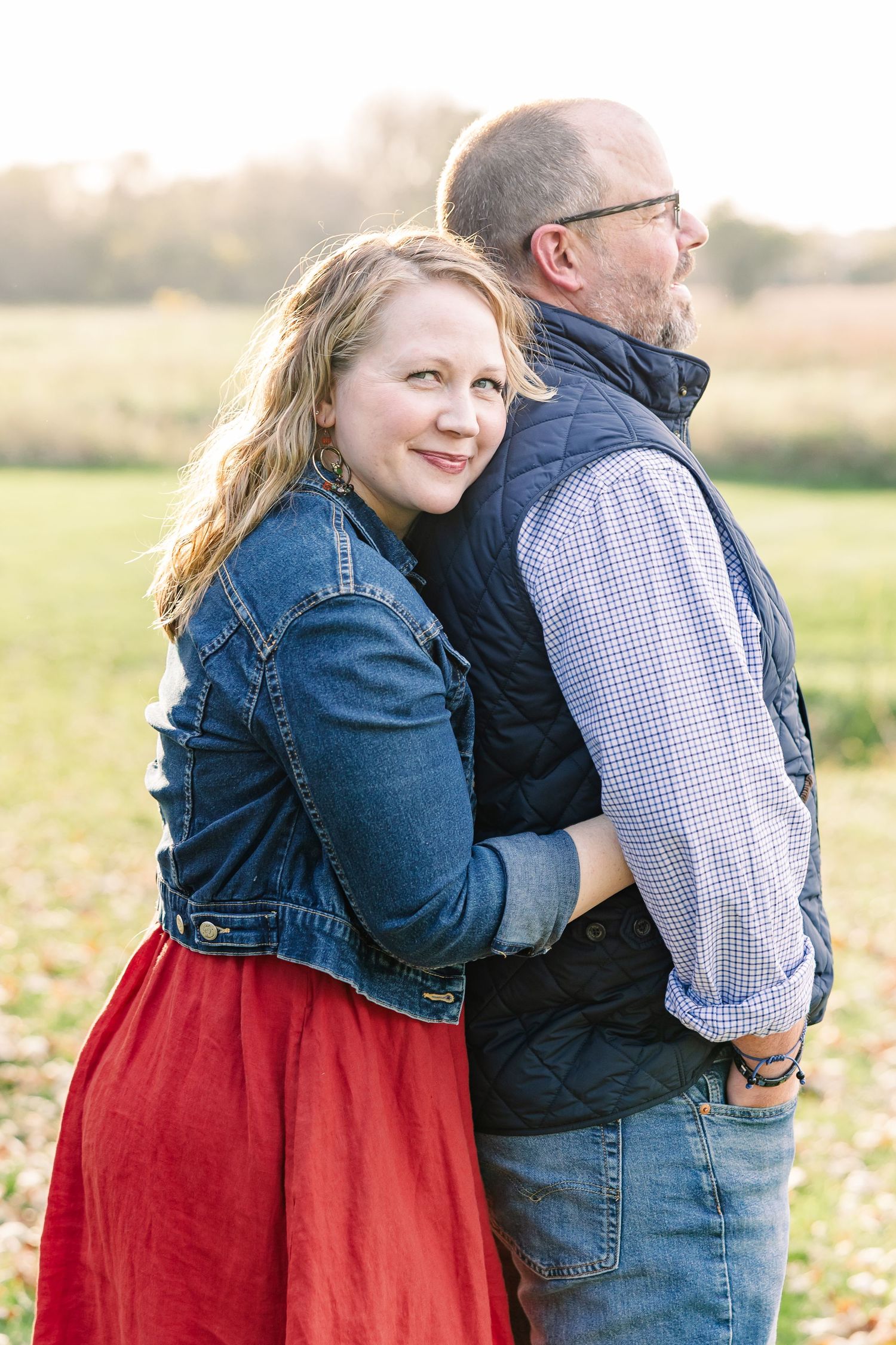 A smiling woman in a denim jacket and red skirt hugs a bearded man in glasses and a quilted vest in a golden autumn field.