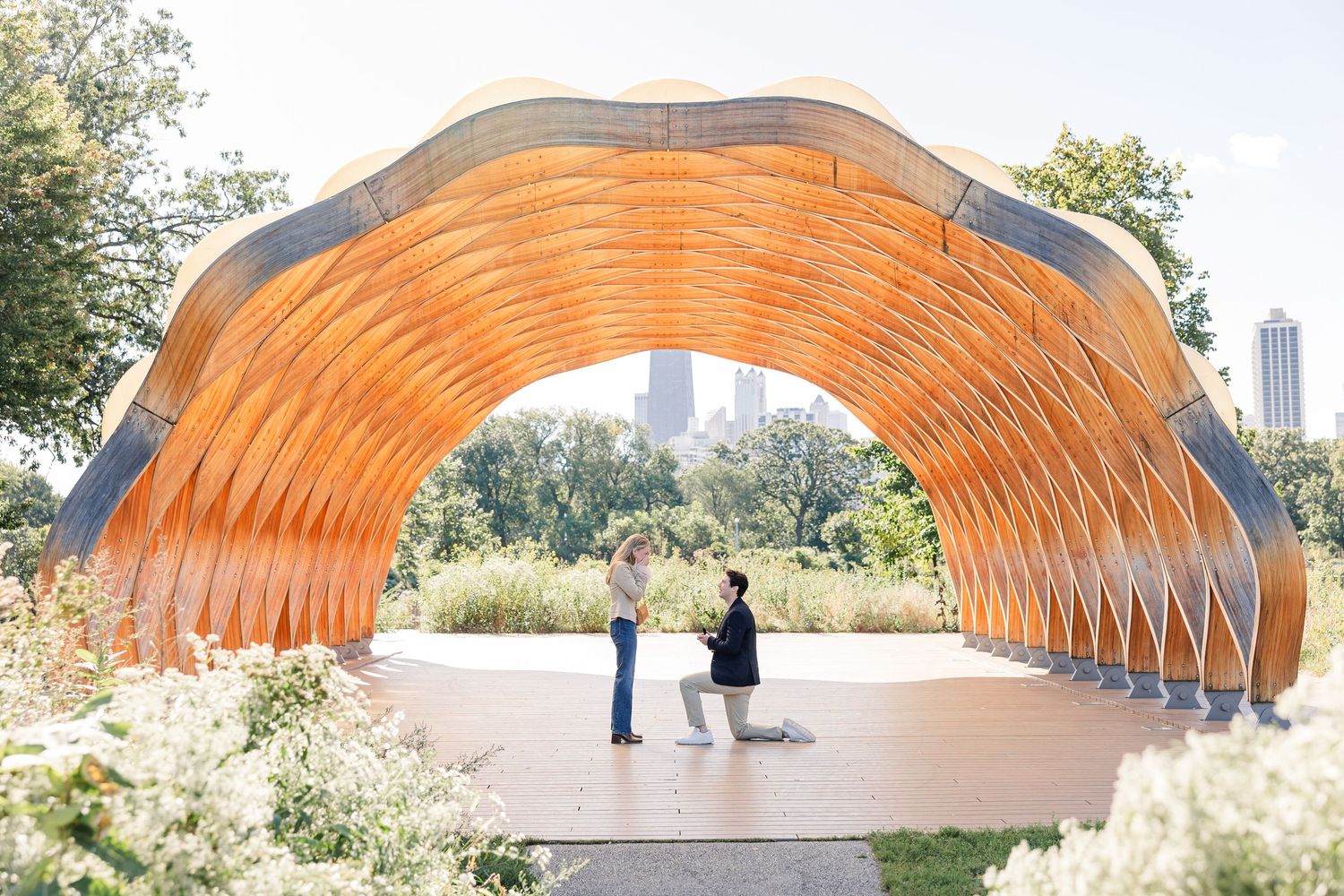 Couple stands under a stunning wooden arch pavilion in a lush park with city skyline in the background.