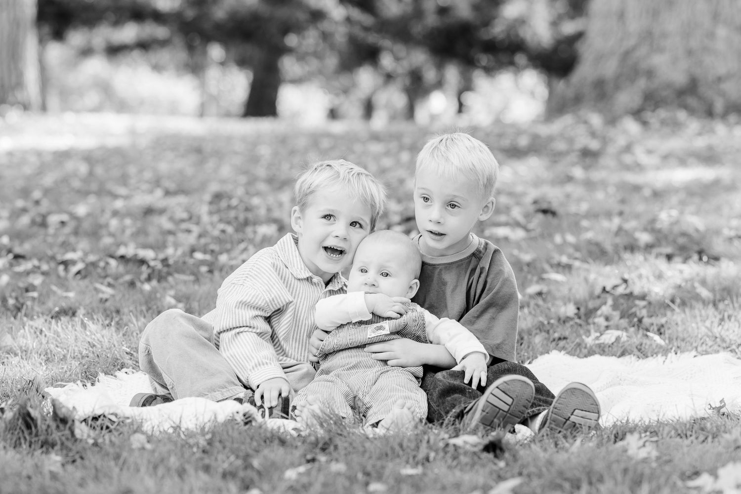 Black and white photo of three young siblings sitting together on grass in a park, smiling warmly.