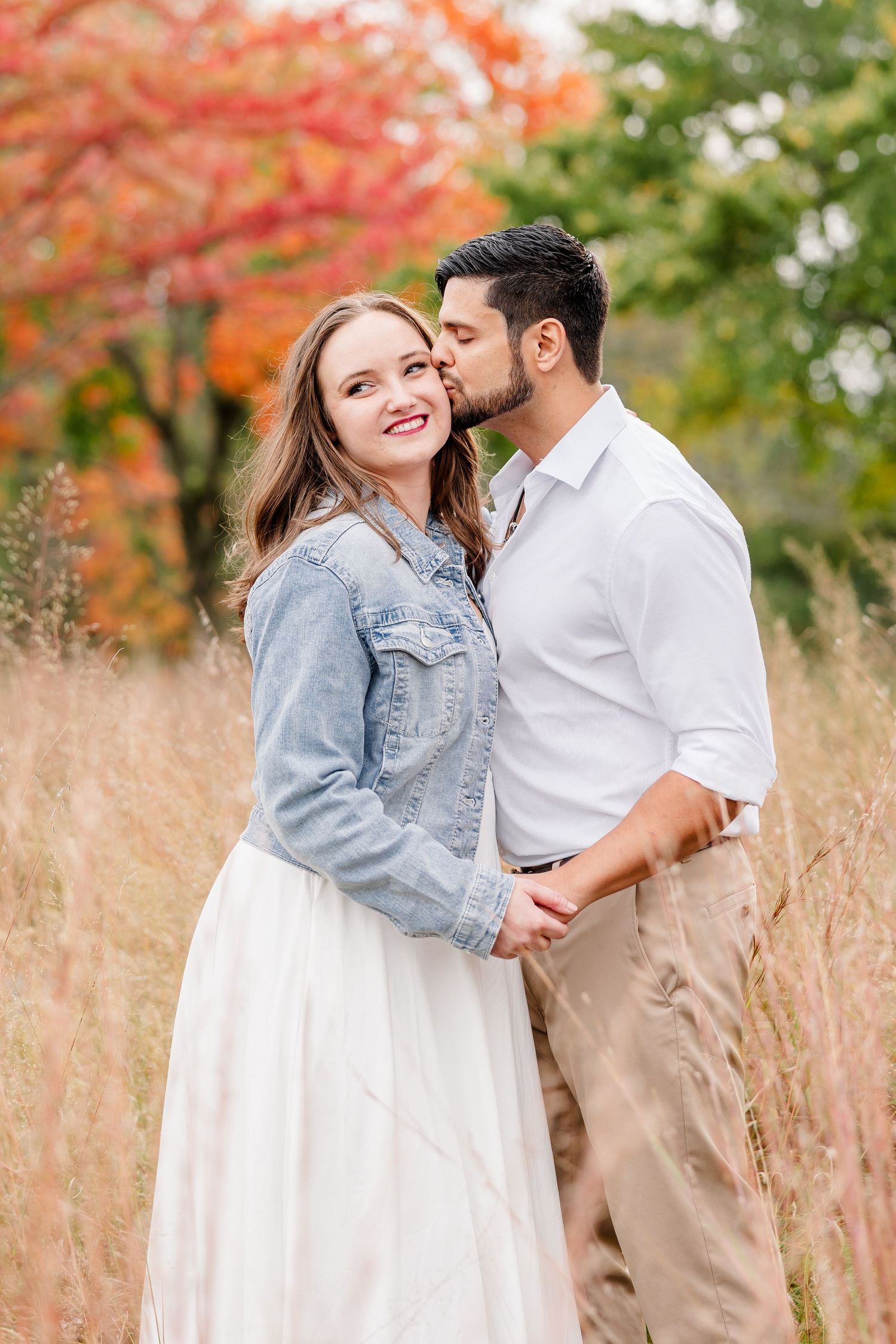 Smiling woman in a denim jacket and white skirt receives a kiss on the cheek from her partner in a golden field with fall trees.