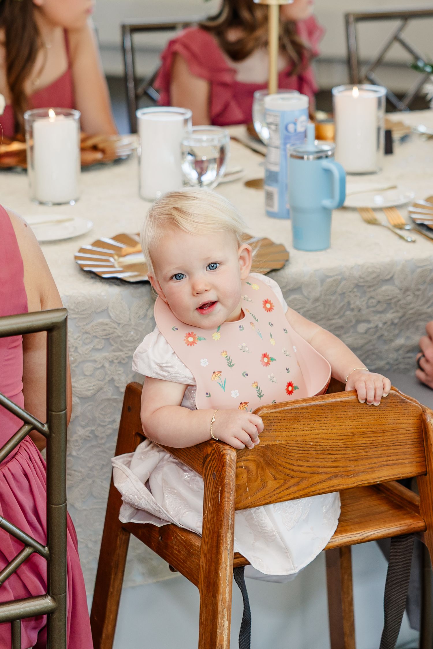 Smiling baby girl in a pink floral bib sits in a wooden high chair at a elegantly set wedding reception table with candles.