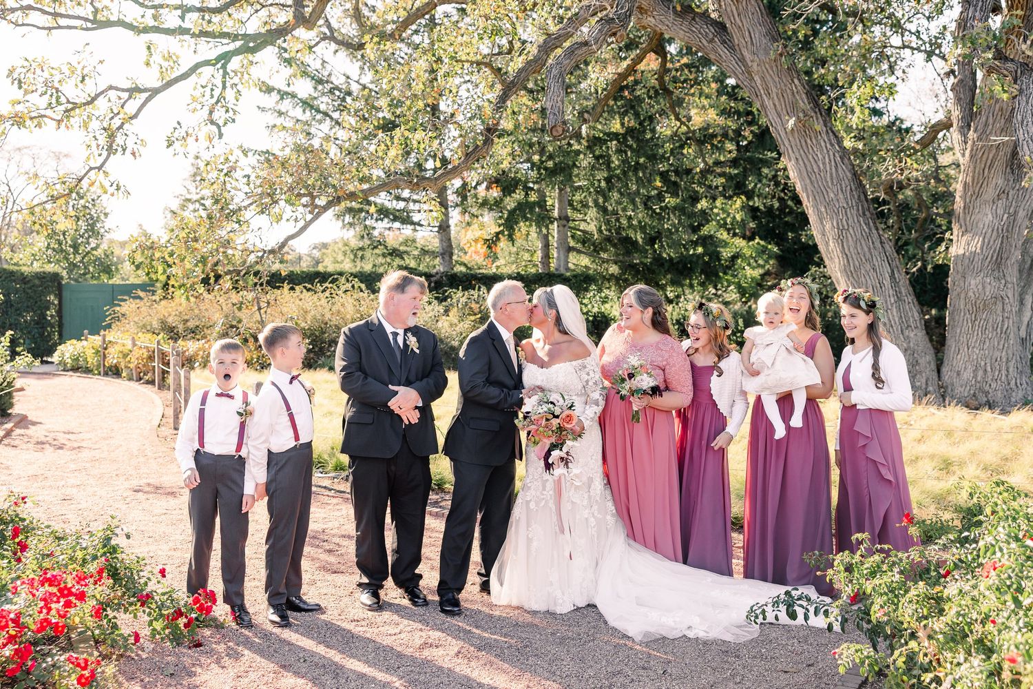 Bride in a white gown holding a bouquet laughs with groom, groomsmen, ring bearers and bridesmaids in plum dresses in a sunny garden.
