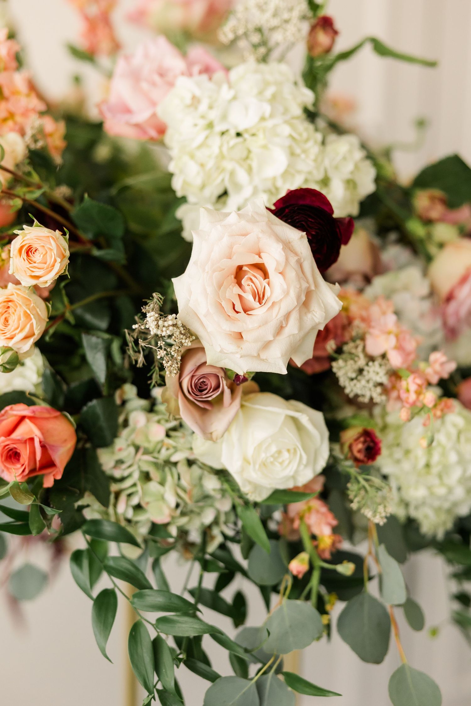 Close-up of a wedding floral arrangement with blush roses, white hydrangea, burgundy blooms, and eucalyptus greenery.