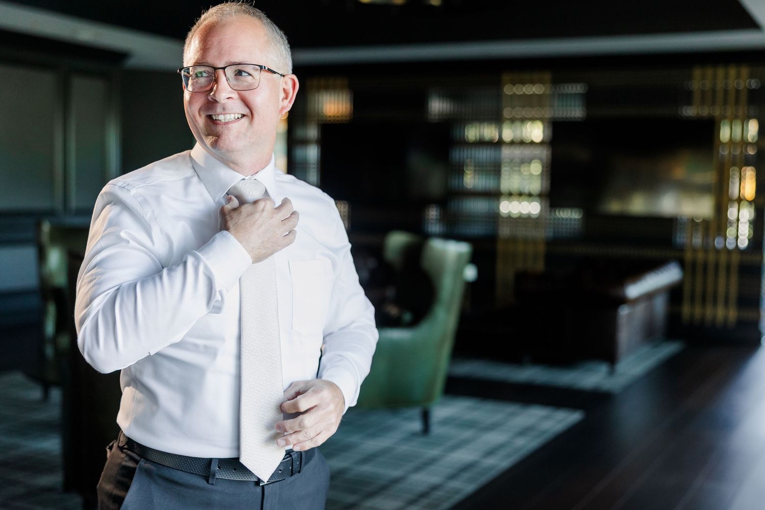 Smiling man in glasses and a white dress shirt adjusts his untied tie in a modern lounge with green armchairs and dark wood decor.