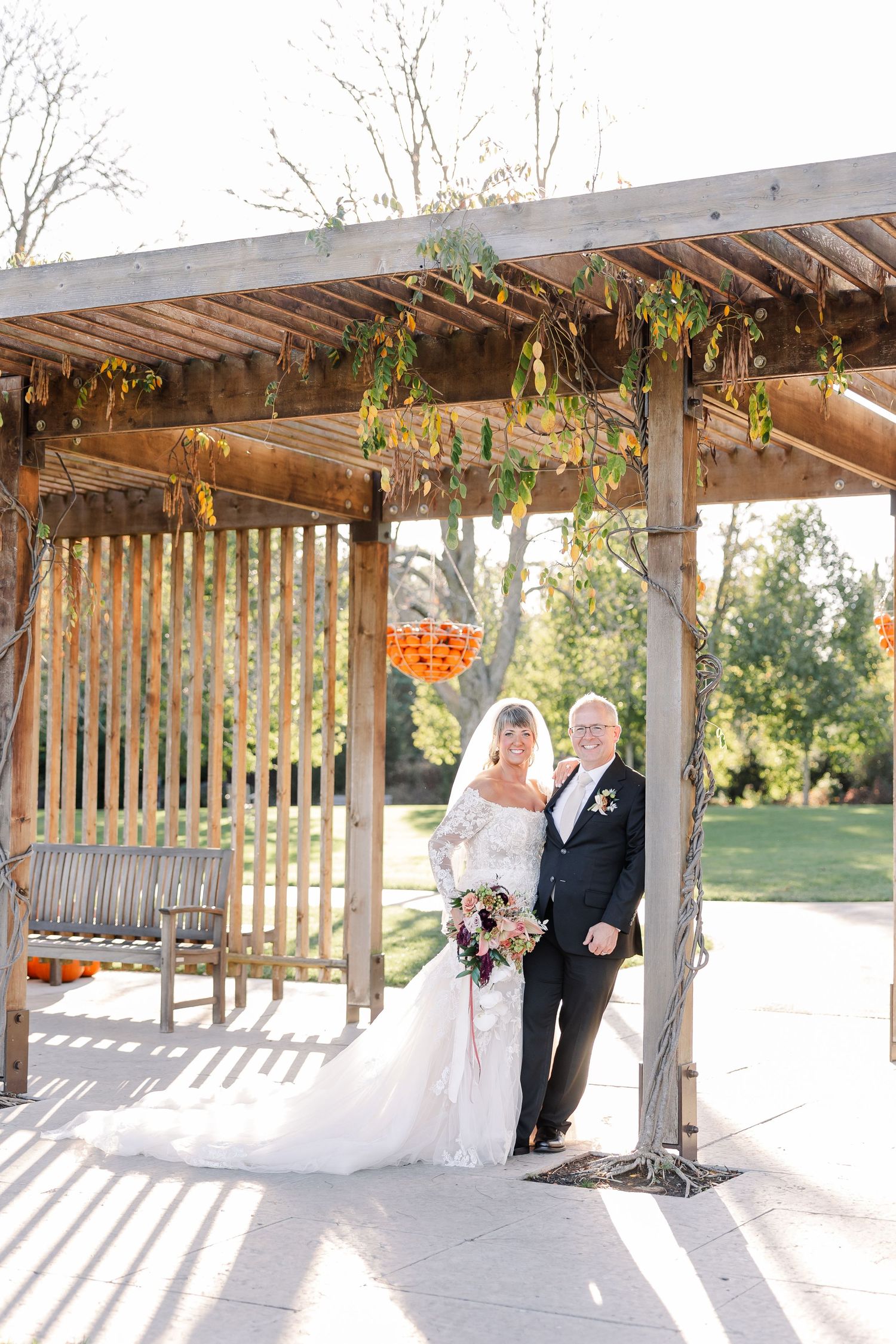 Bride in a lace wedding gown holding a bouquet poses with a groom in a dark suit beneath a vine-covered wooden pergola in autumn.
