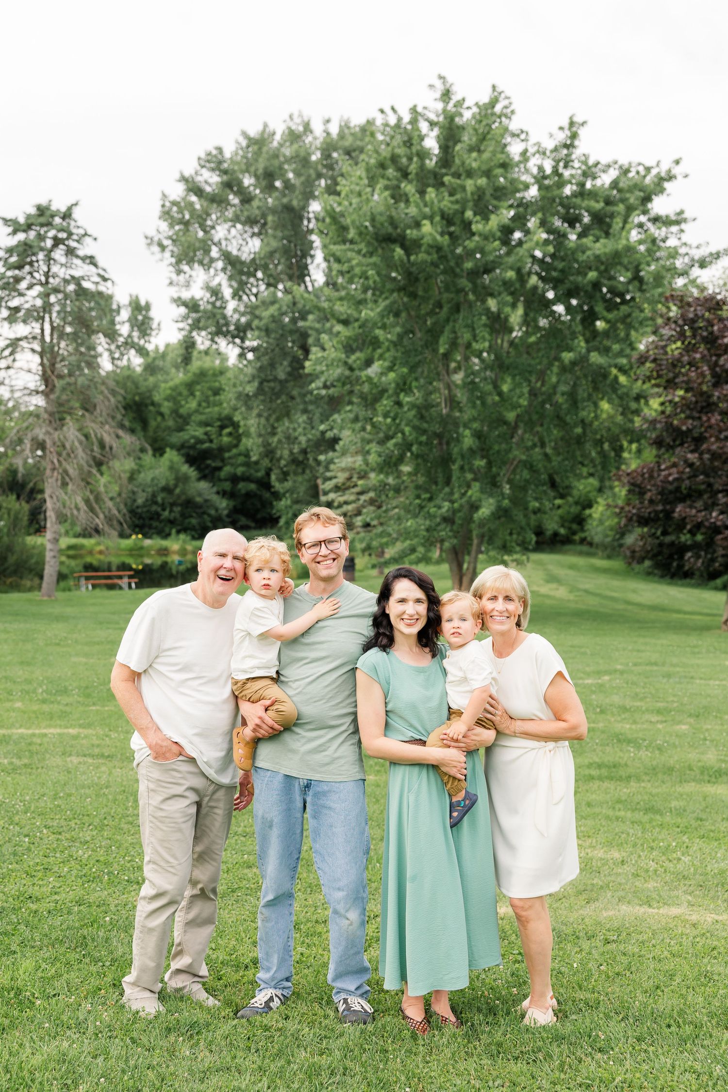Multi-generational family of six smiling together on a green lawn with trees behind them.
