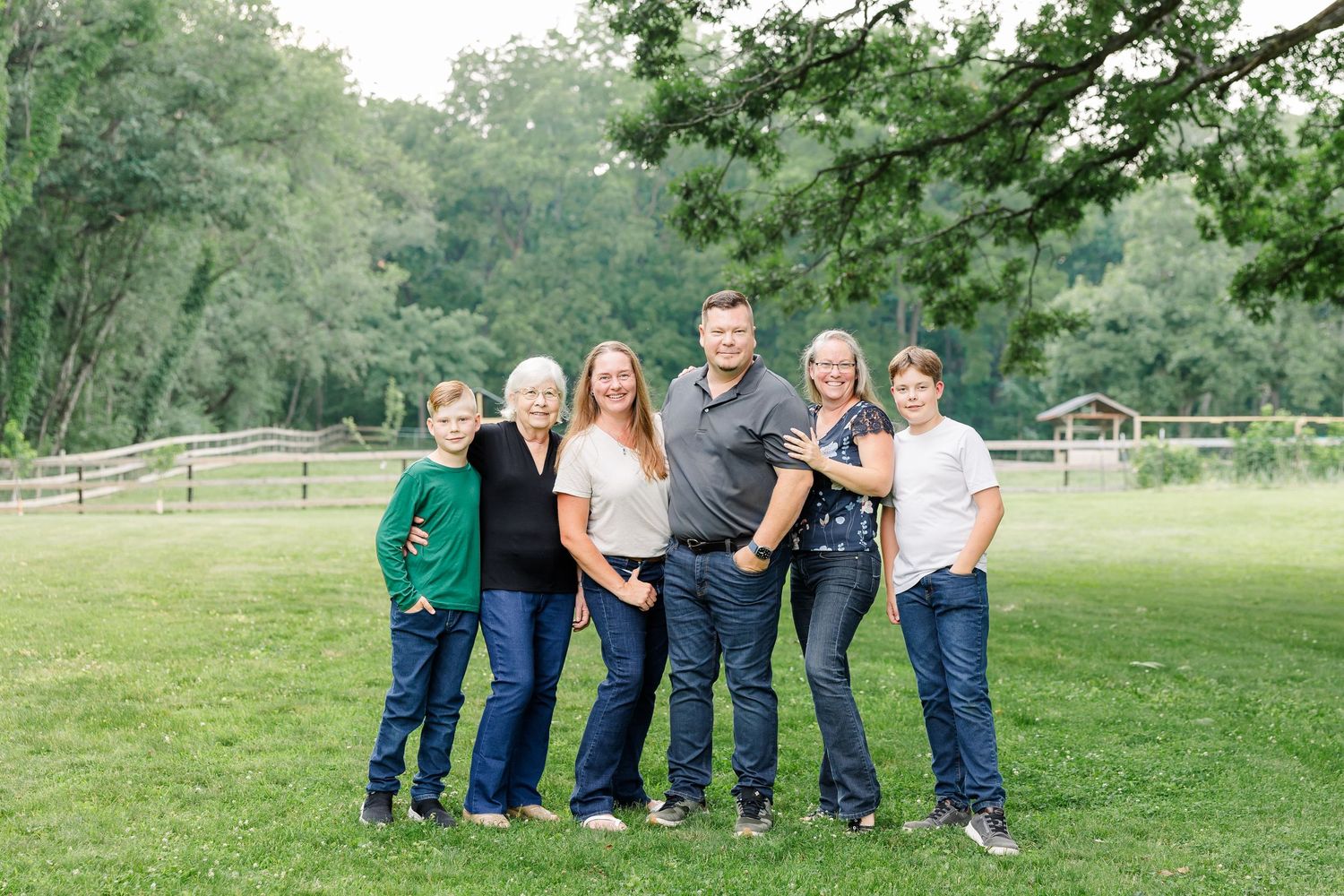 Family of six posing together outdoors on green grass with trees and wooden fence in background.