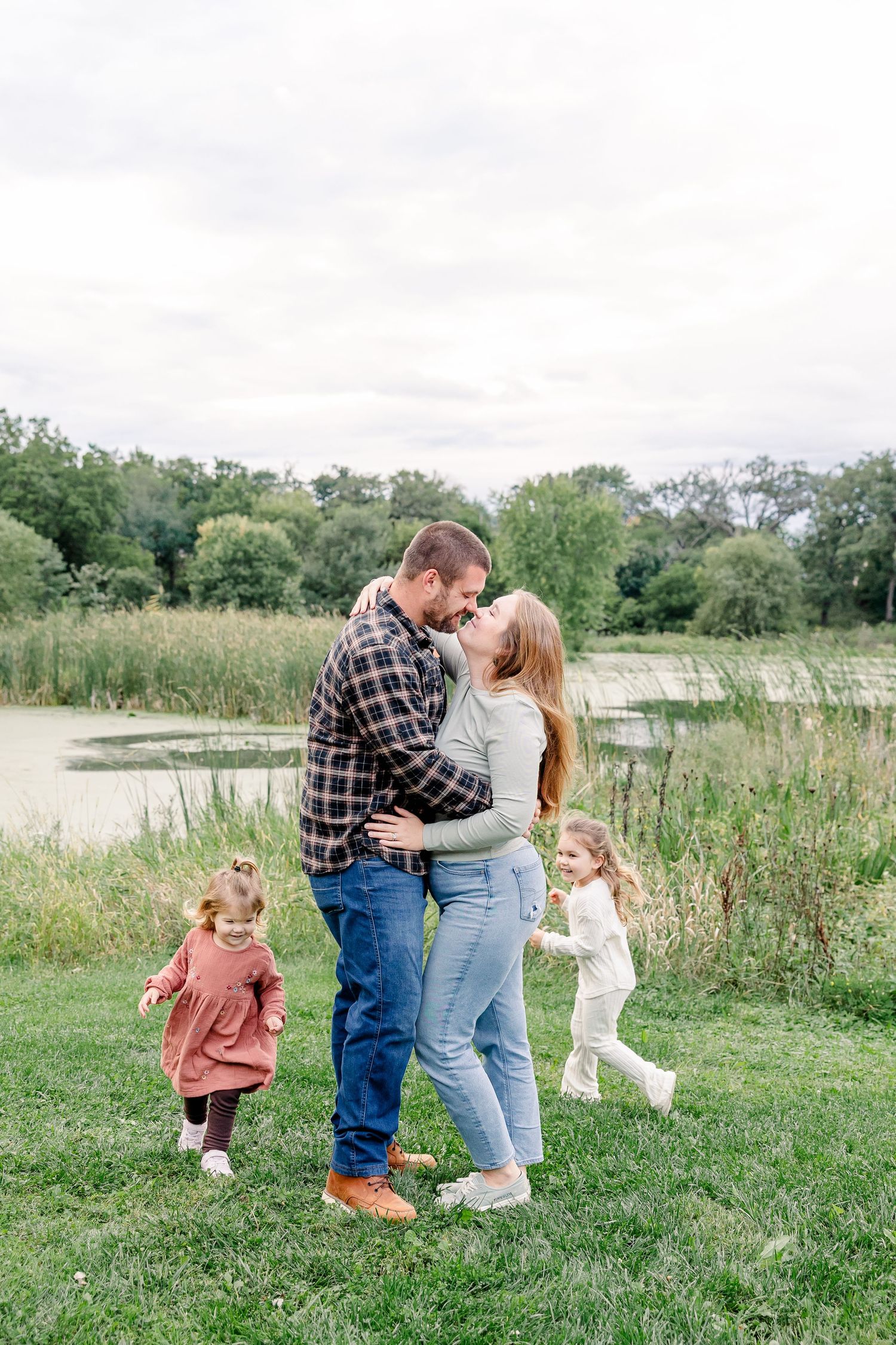 A loving couple kisses near a pond while their two young daughters play around them on green grass.