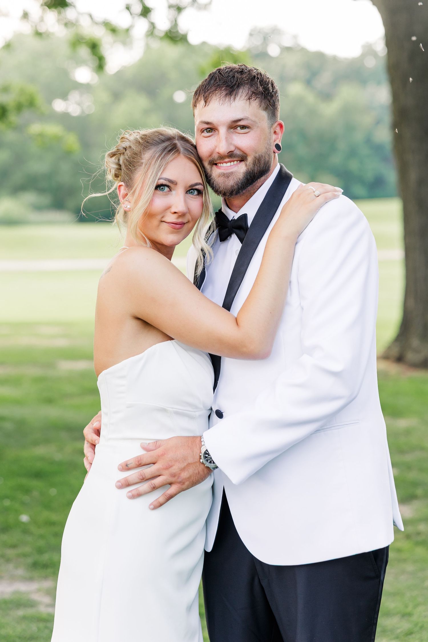 Smiling bride and groom embrace outdoors, groom wearing a white tuxedo jacket and black bow tie.