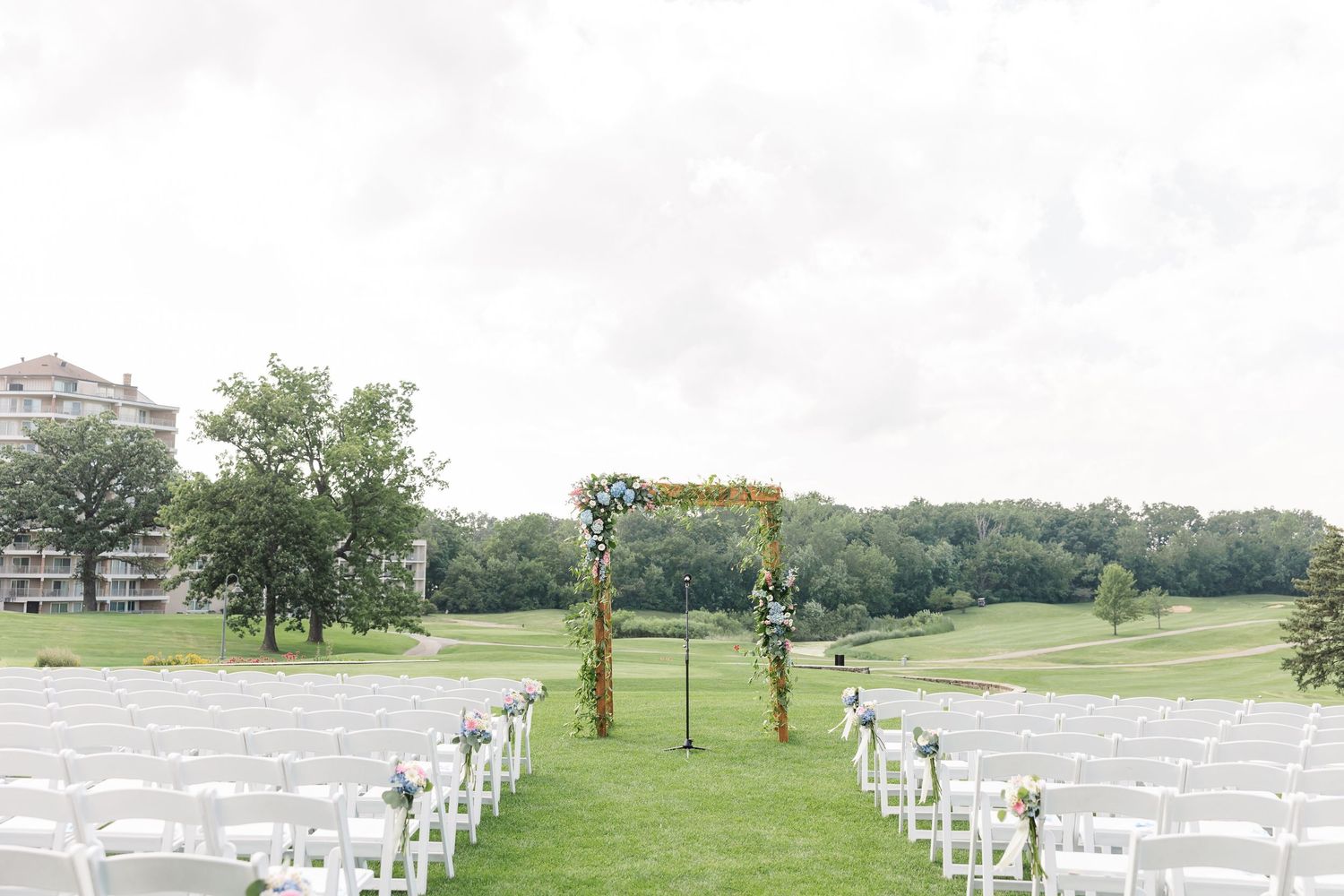 Elegant outdoor wedding ceremony setup with white chairs, floral arch, and lush green golf course backdrop.