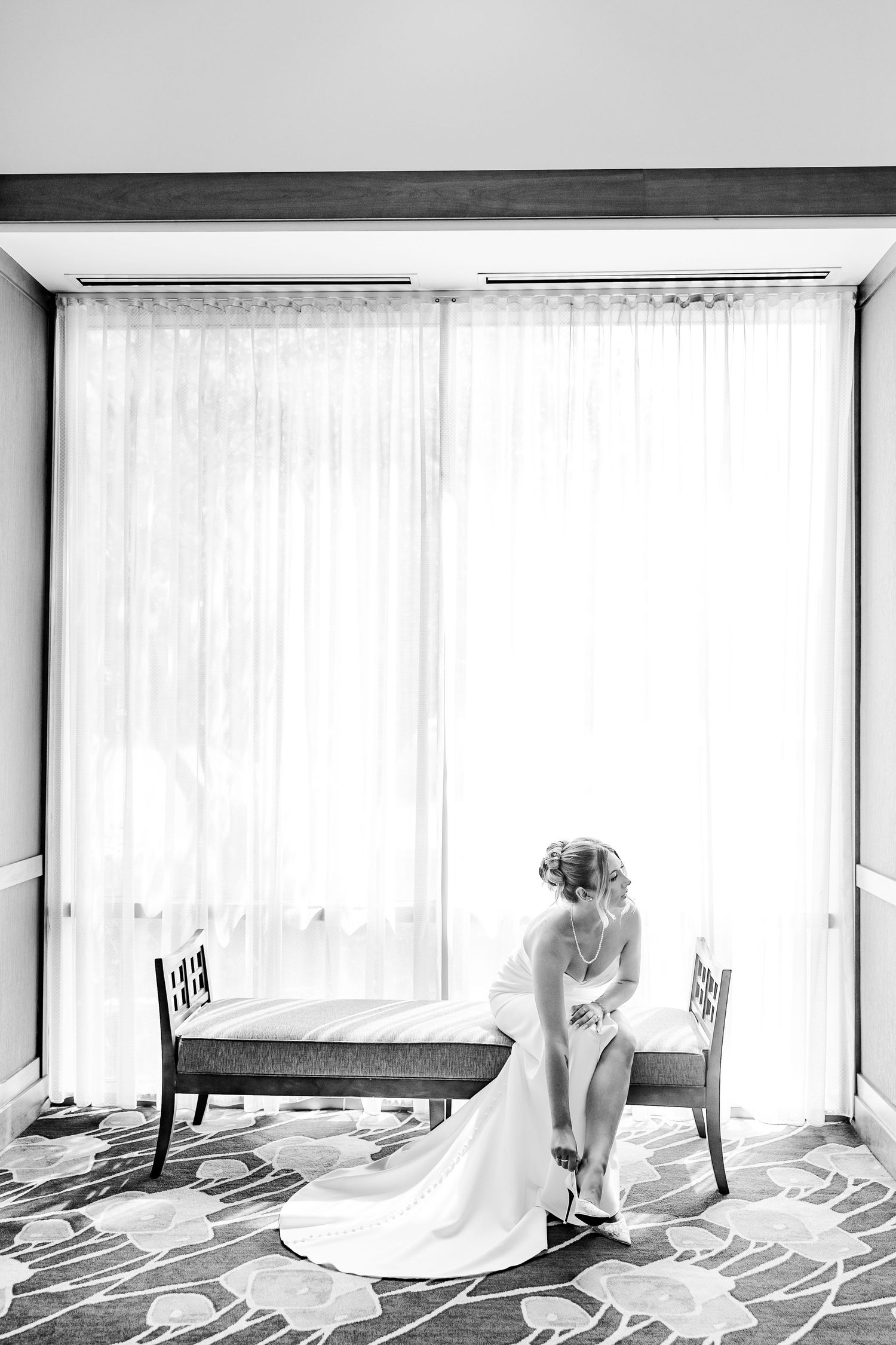 Bride in white gown sits on bench adjusting her shoes in black and white wedding photo.