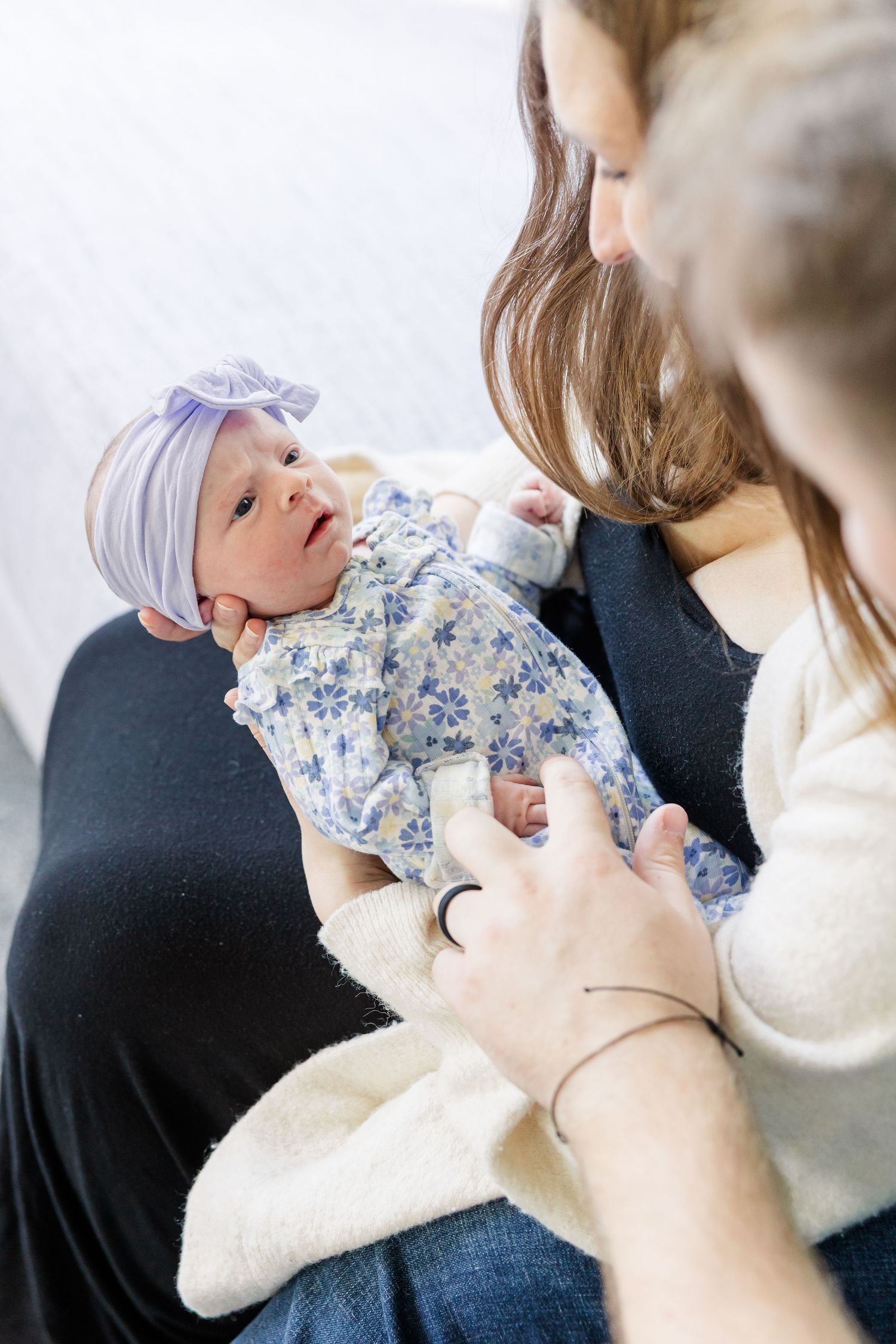 A mother tenderly holds her newborn baby girl dressed in a floral outfit with a purple bow headband.