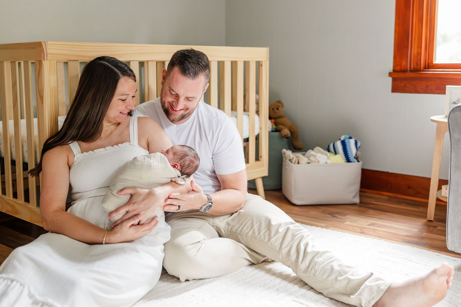 New parents sitting on floor by crib, lovingly gazing at their newborn baby in a cozy nursery room.