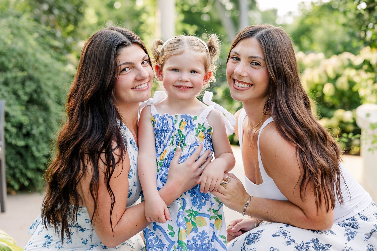 Two young women in floral dresses smiling with a little girl in a blue floral dress outdoors.