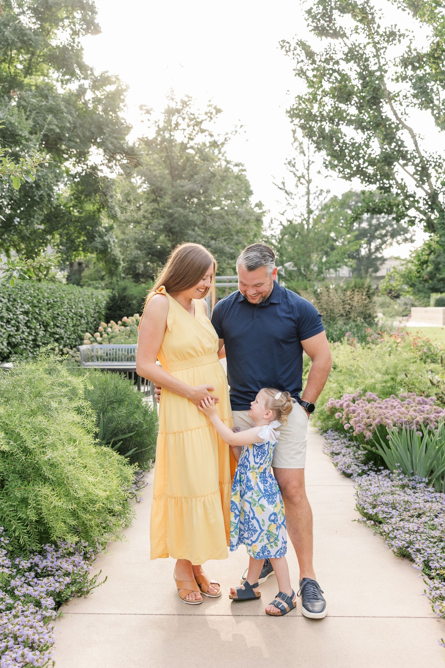Pregnant woman in yellow dress with husband and toddler daughter in blue floral dress in a garden.