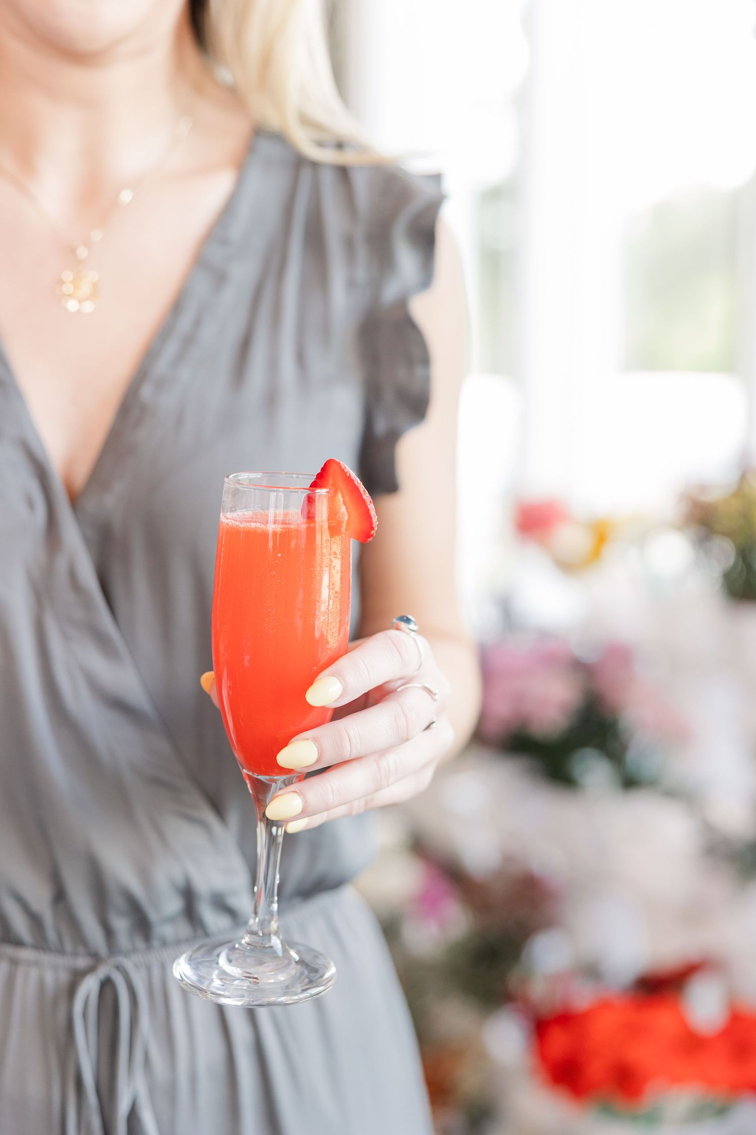 Woman in gray wrap dress holding a strawberry mimosa cocktail in a champagne flute at a brunch event.