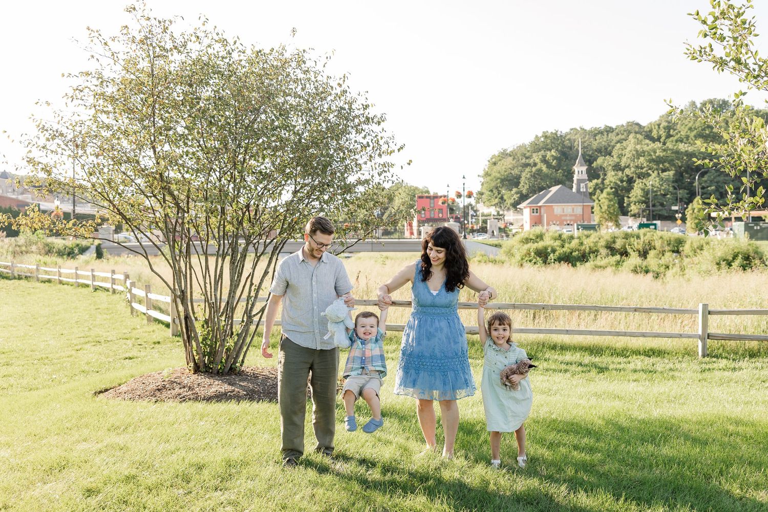 Family of four walking on green grass near a flowering shrub on a sunny day in a charming small town.