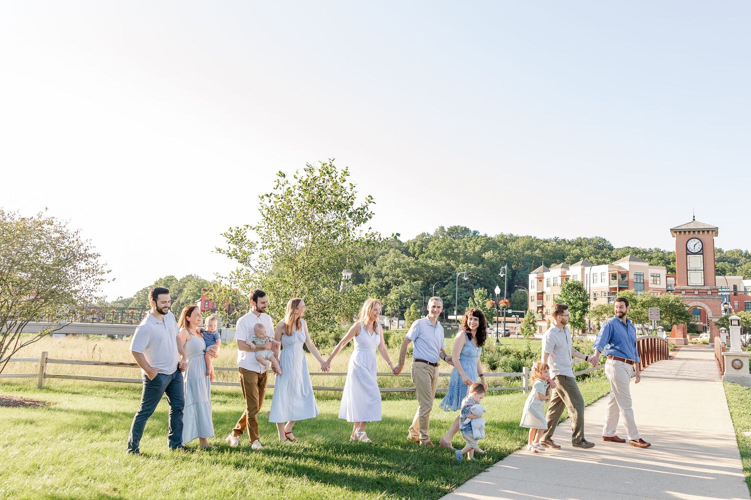 An extended family walks together along a sunny outdoor path surrounded by green grass and trees.
