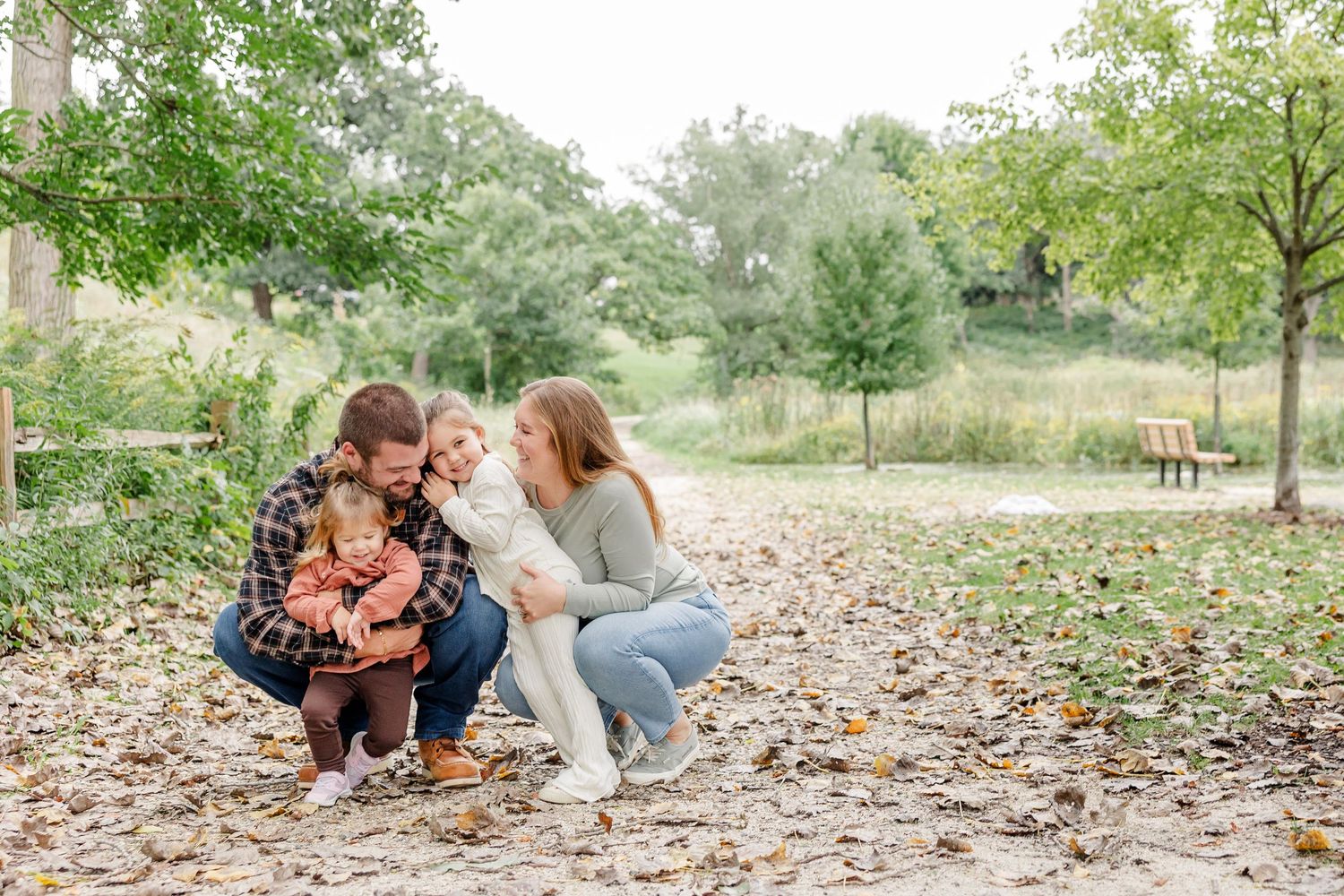 Family of four sharing a warm embrace outdoors among fallen autumn leaves in a lush green park setting.