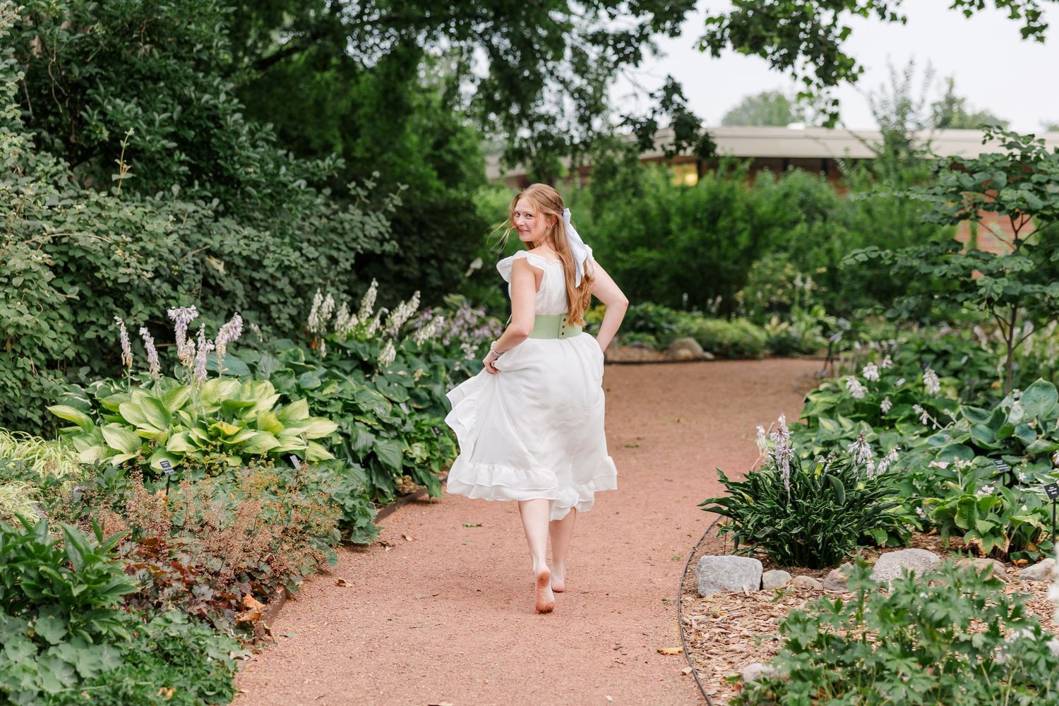 A woman in a white dress twirls on a garden path surrounded by lush green plants and flowers.