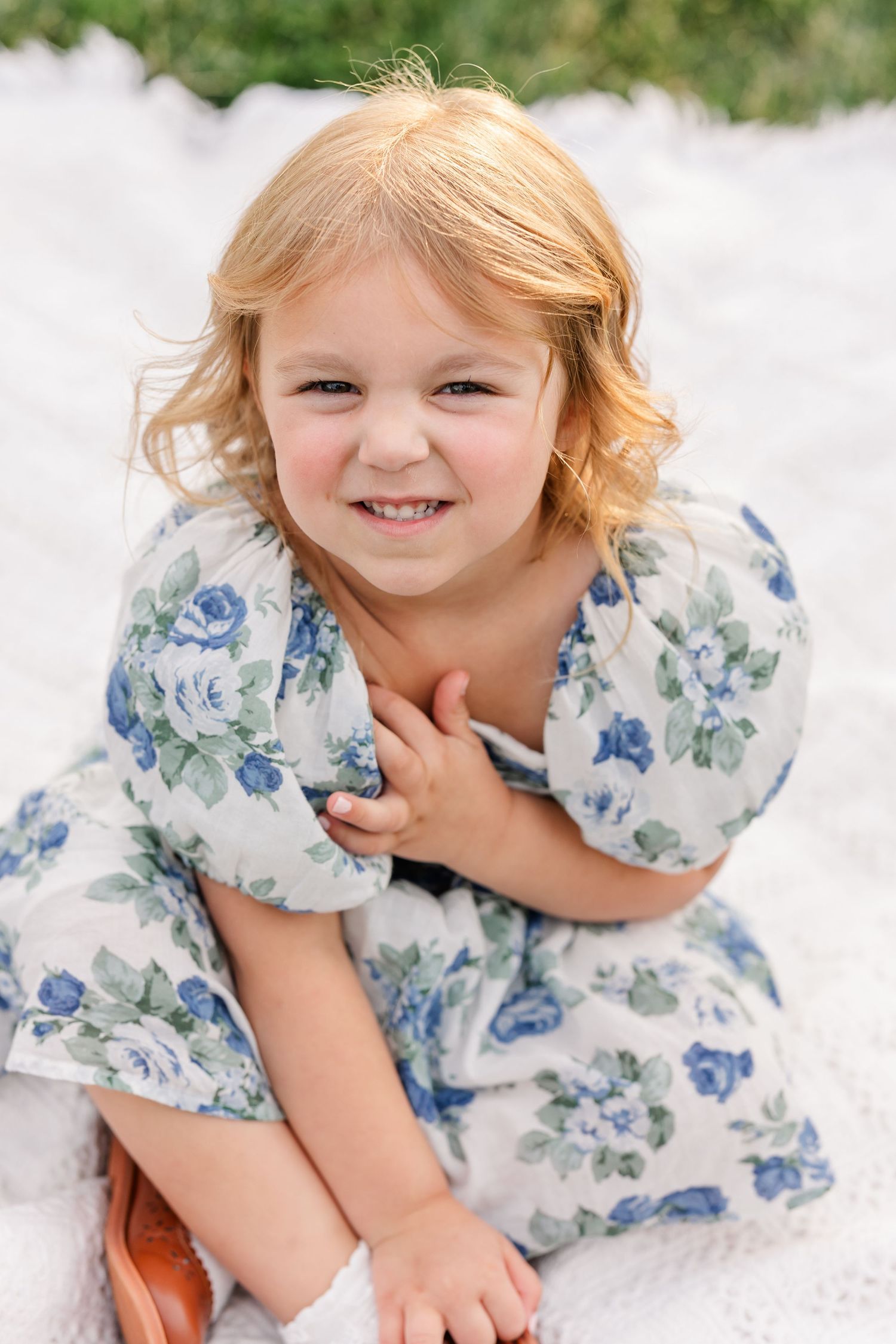 Smiling young girl with blonde hair wearing a blue floral dress sits on a white blanket outdoors.