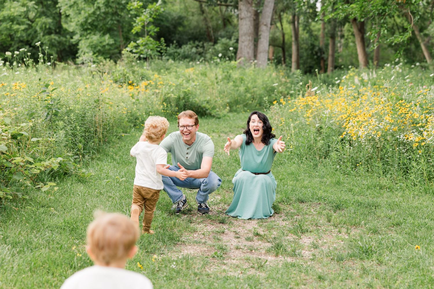 A joyful family with two young children plays in a lush green meadow with wildflowers during an outdoor photo session.