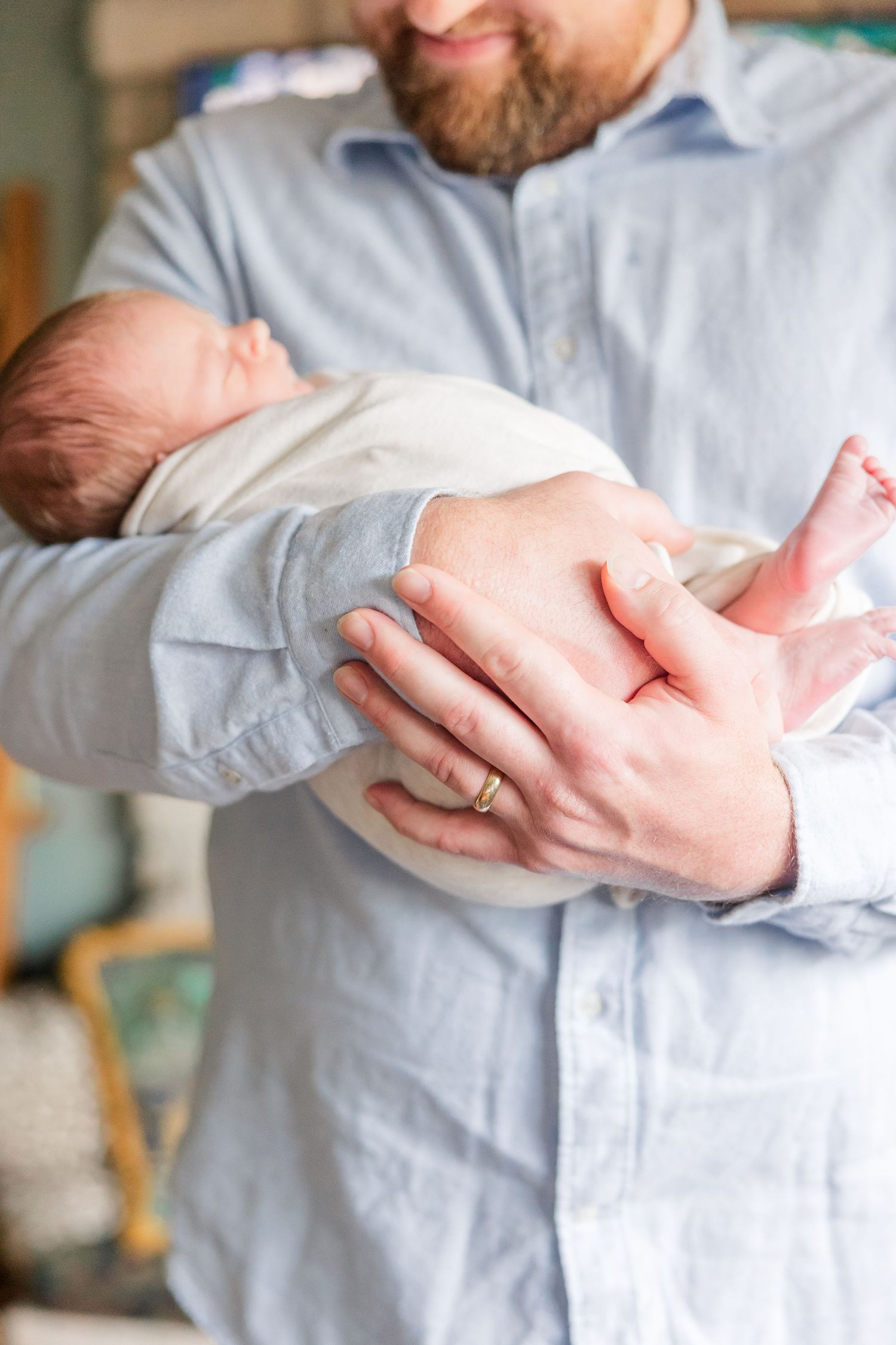 Bearded father in blue shirt cradling swaddled newborn baby in his arms, smiling warmly.
