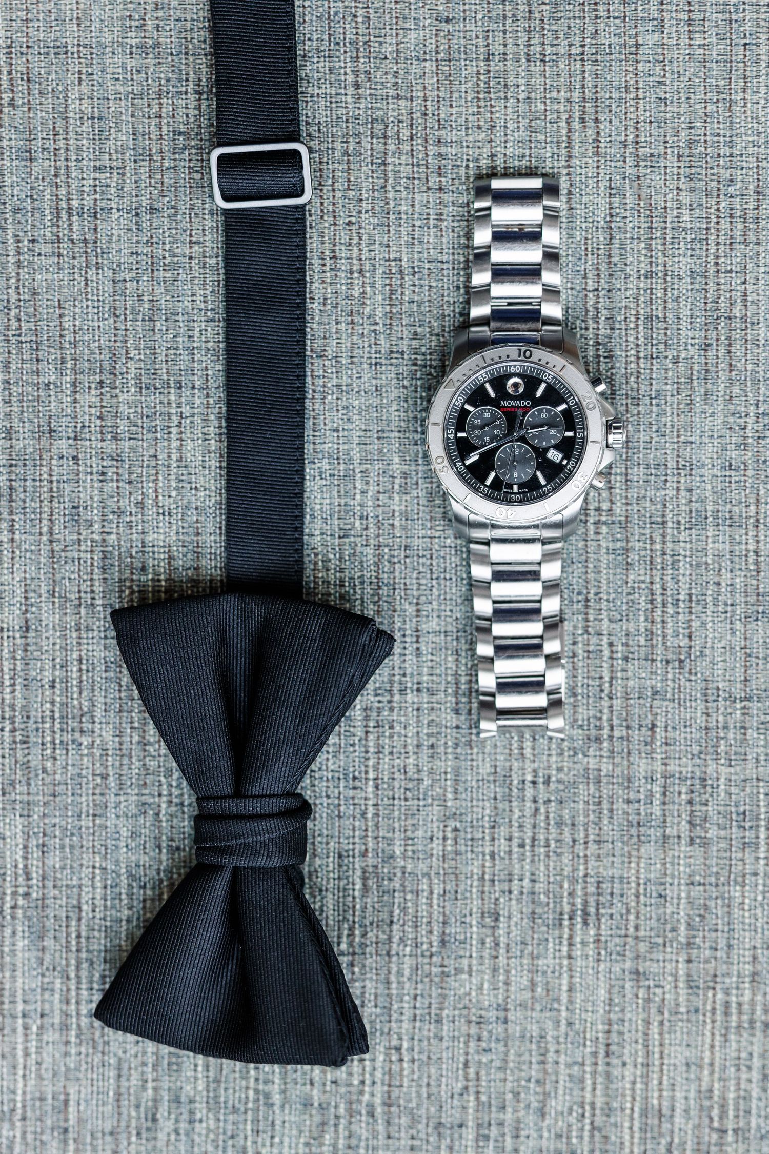Black bow tie and silver chronograph watch laid flat on gray textured surface.