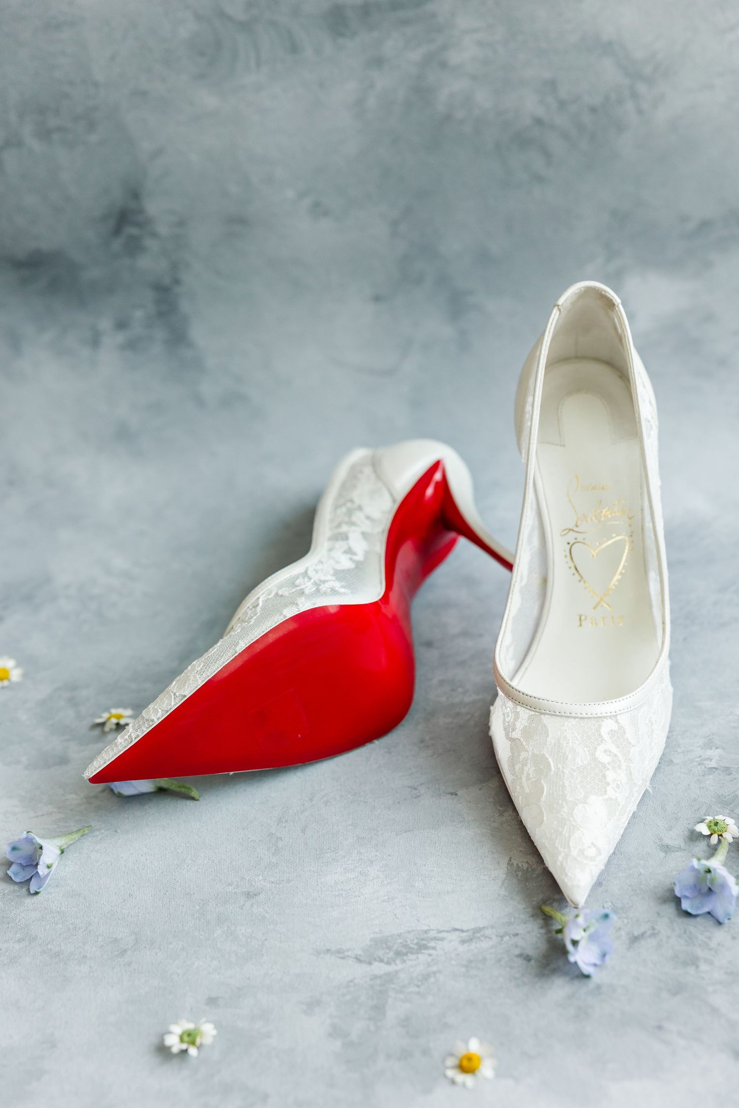 White lace bridal heels with red soles displayed on gray surface with delicate blue and white flowers.