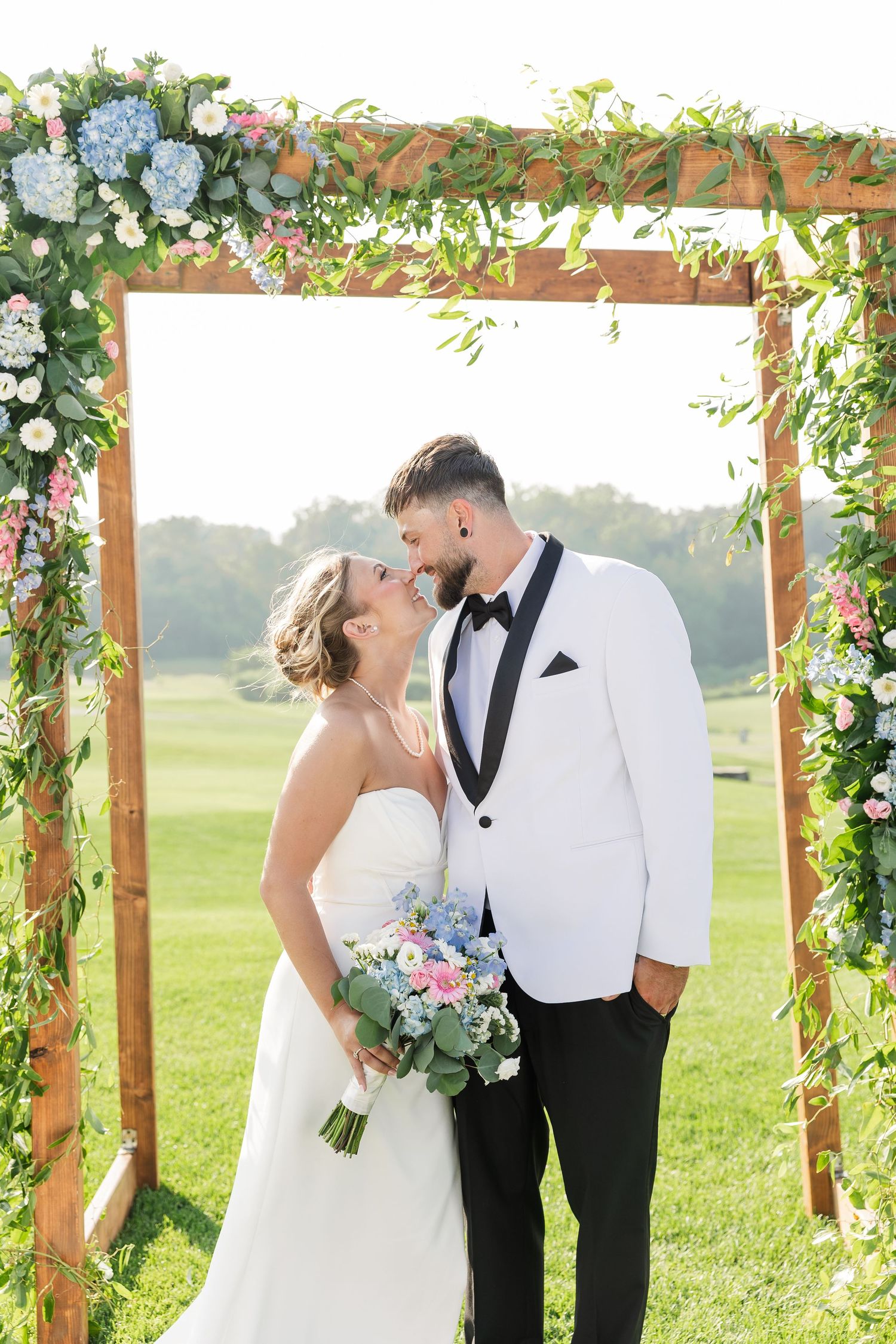 Bride and groom kissing under floral wooden arch decorated with hydrangeas and greenery.