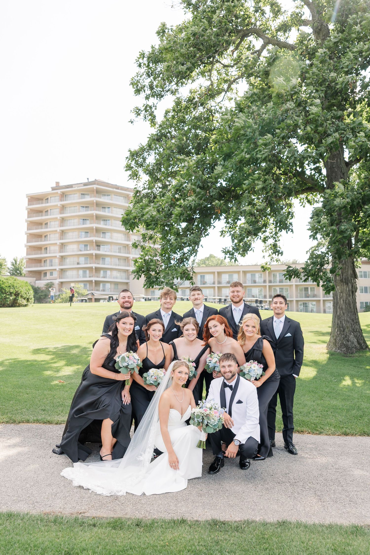 Wedding party poses outdoors on a golf course, bridesmaids in black gowns and groomsmen in dark suits with a building backdrop.