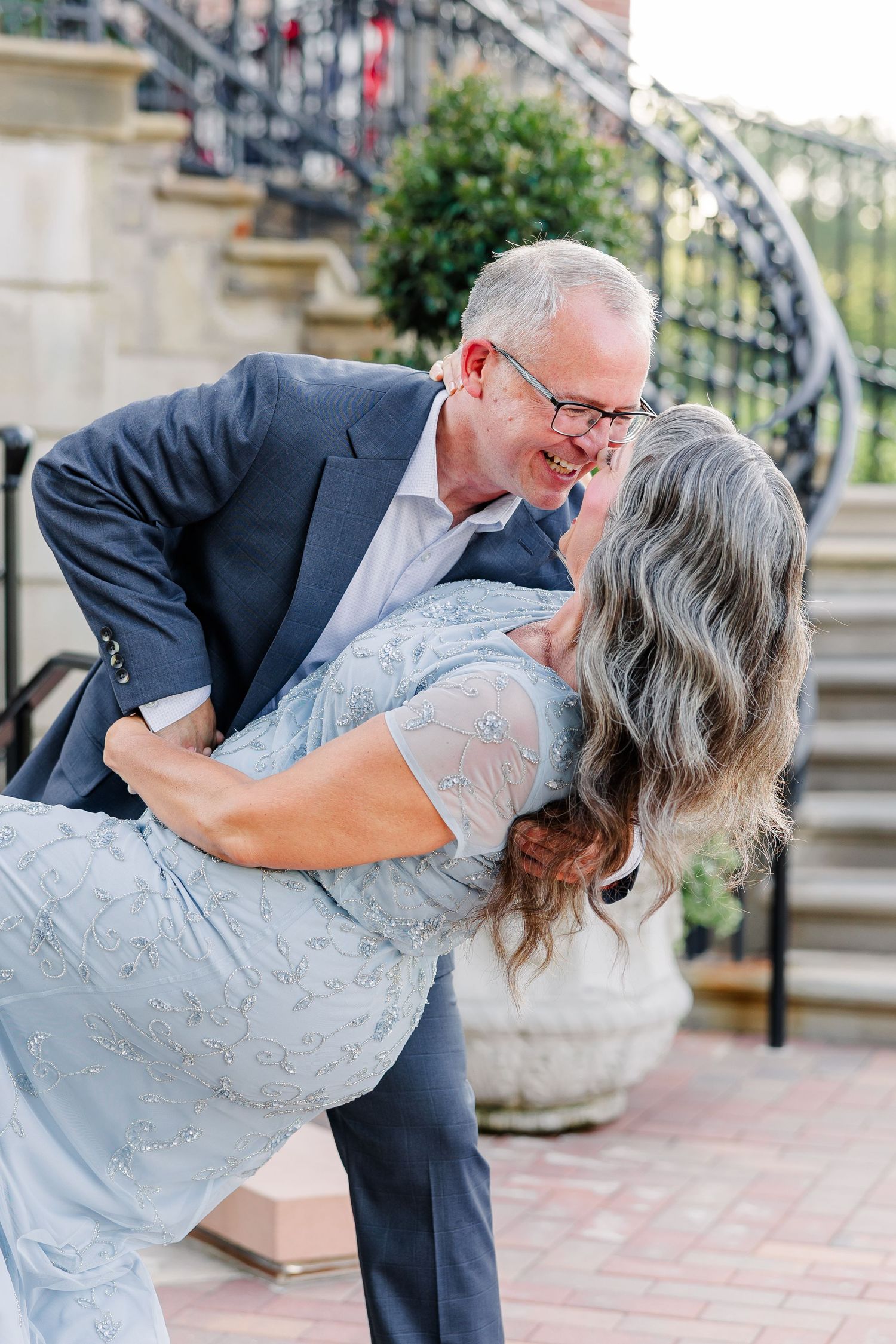 A joyful older couple shares a romantic dip and laugh together on a brick patio near elegant iron stairs.