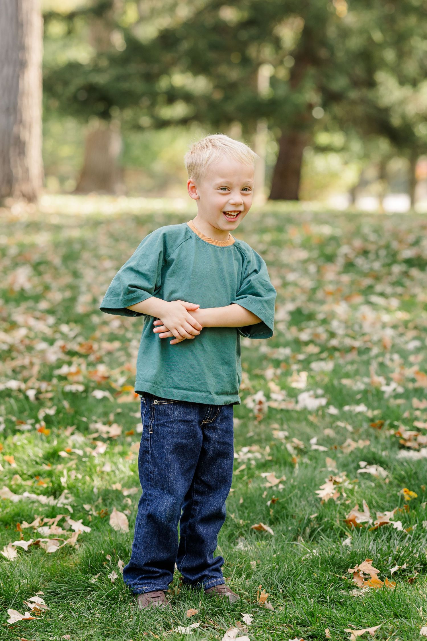 Happy blonde toddler boy in green shirt laughing while standing on grass with fallen leaves.