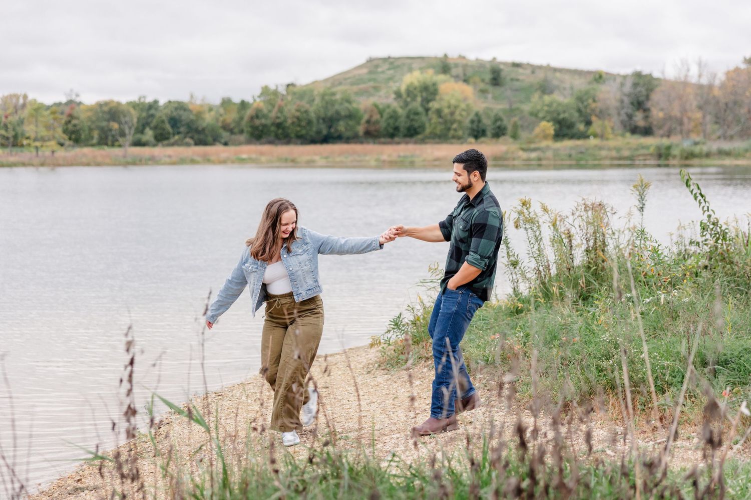 Couple holding hands and walking along a grassy lakeshore with rolling hills and fall foliage in the background.