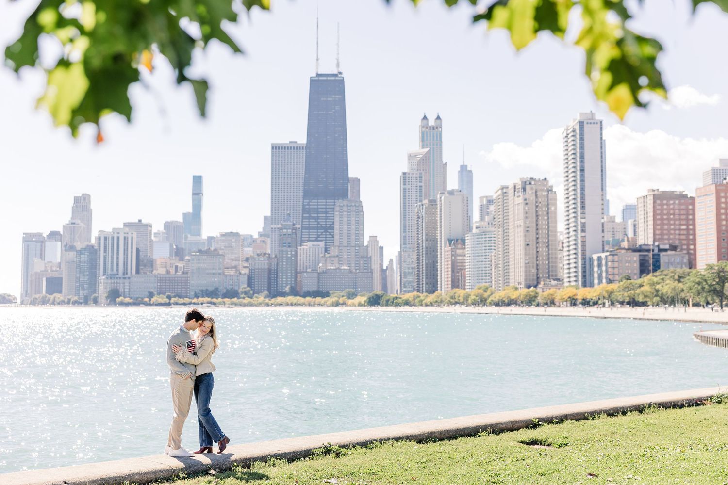 Couple embracing by Lake Michigan with Chicago skyline and John Hancock Center in background on sunny day.