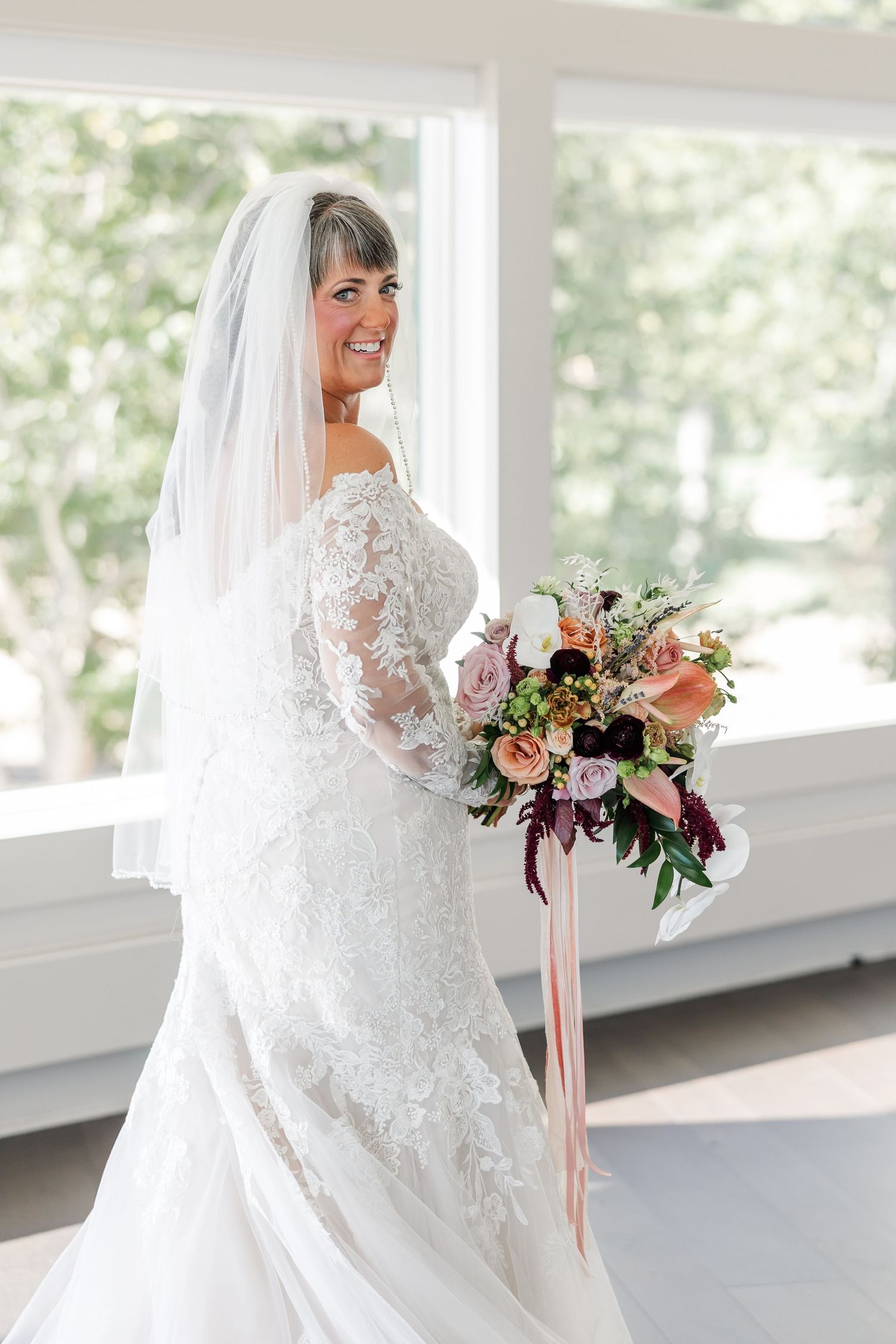 Smiling bride in a lace off-the-shoulder wedding gown and long veil holds a colorful bouquet with trailing ribbons by a bright window.