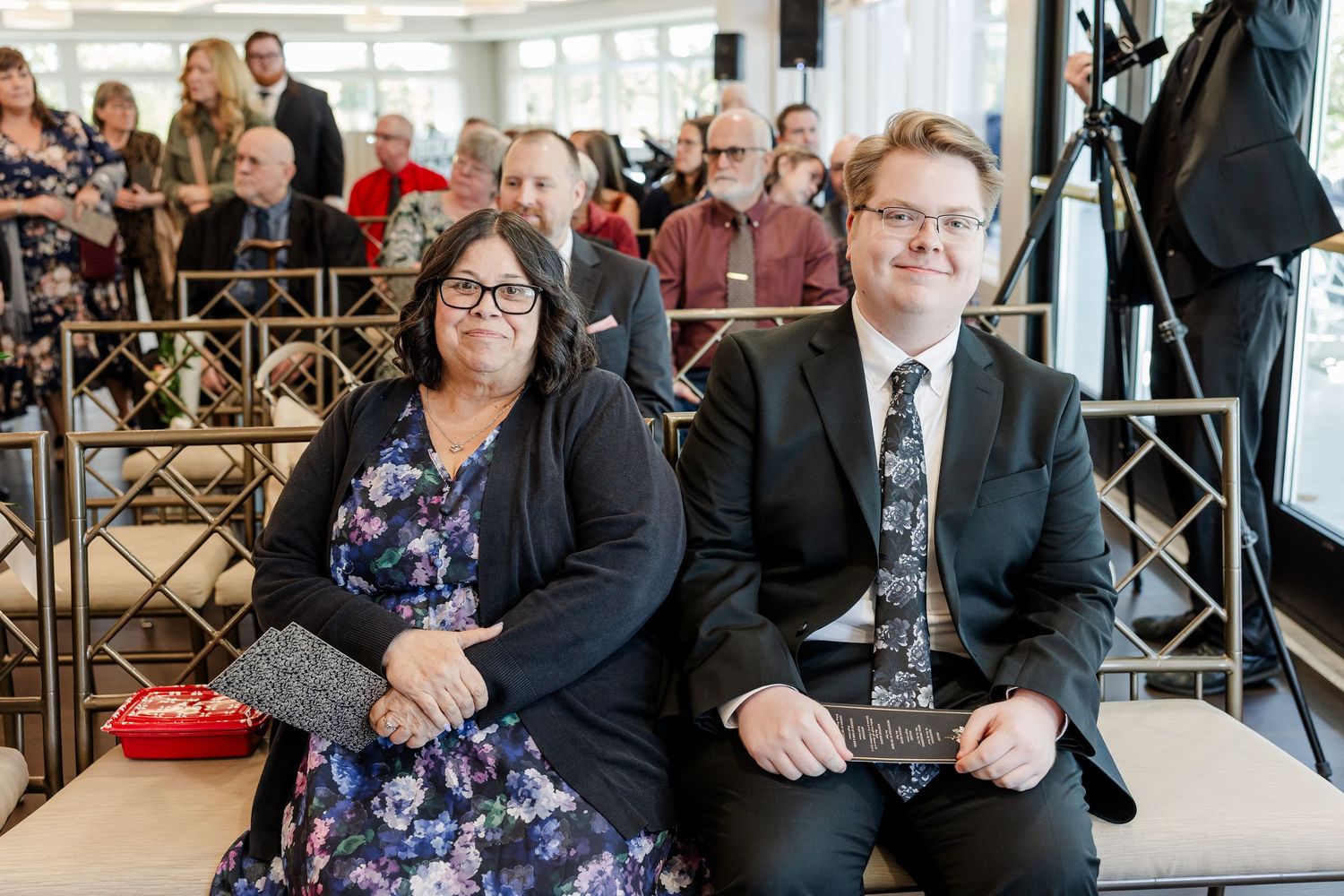 Woman in a floral dress and cardigan sits beside a man in a dark suit and tie at an indoor event with seated guests behind them.