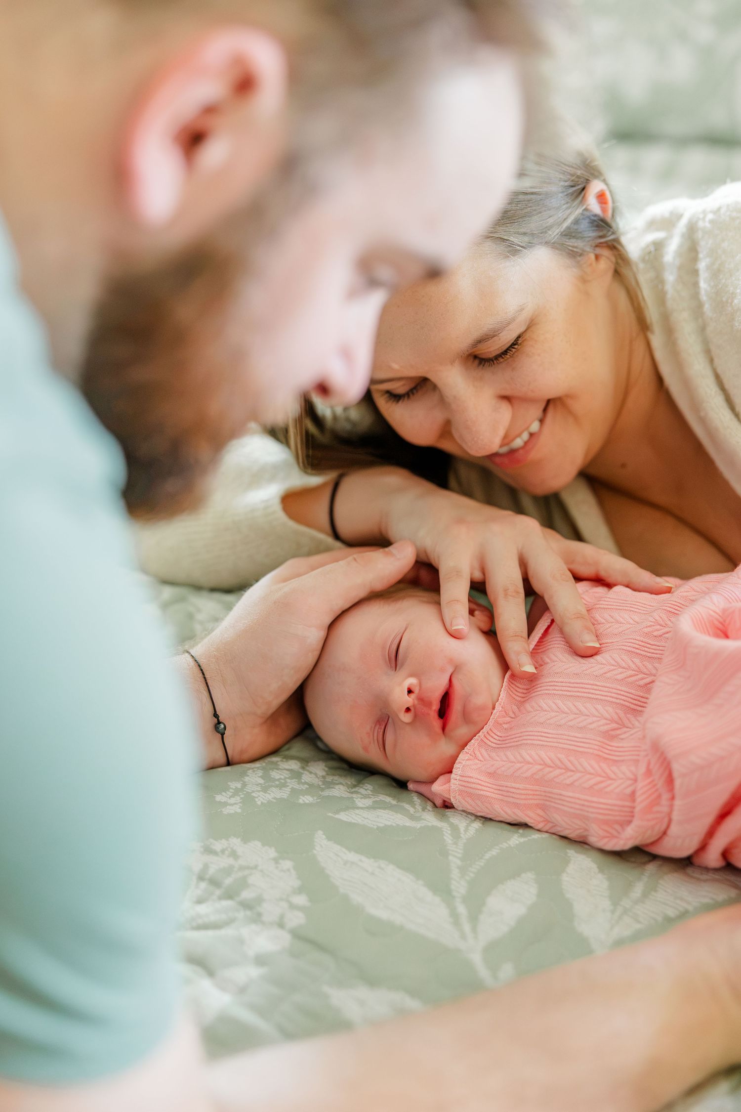 Parents lovingly gazing at their sleeping newborn baby dressed in pink on a floral green bed.