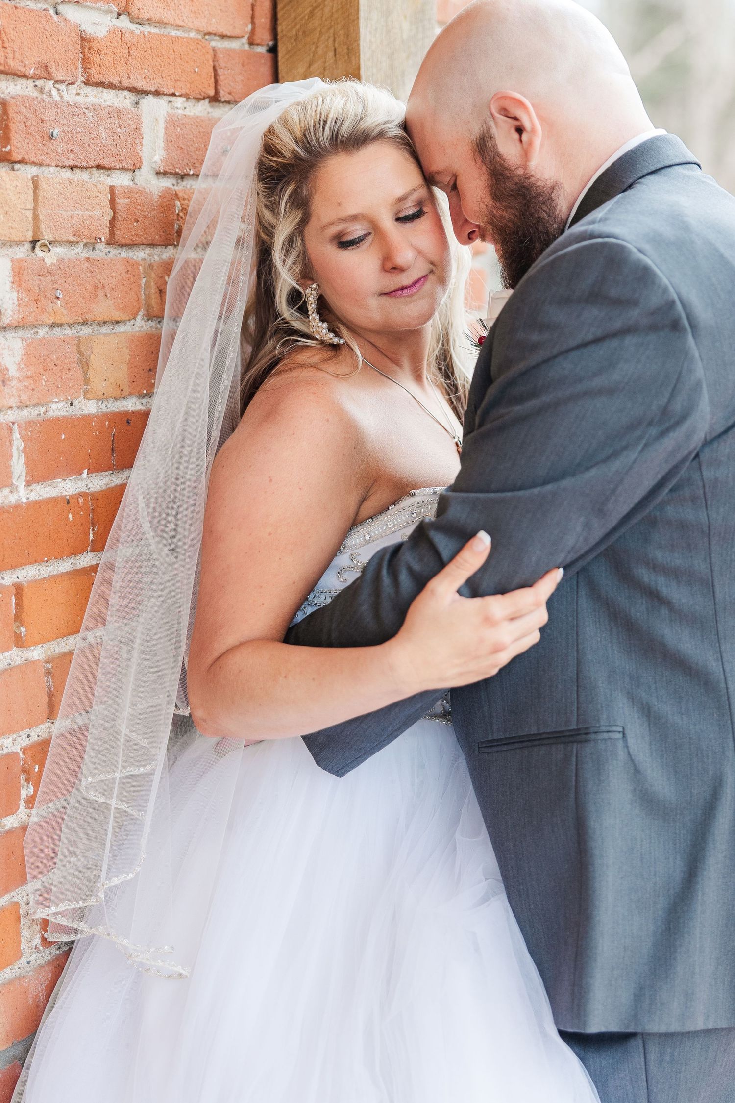 A romantic wedding moment captured against a brick wall as the couple embraces in formal attire.