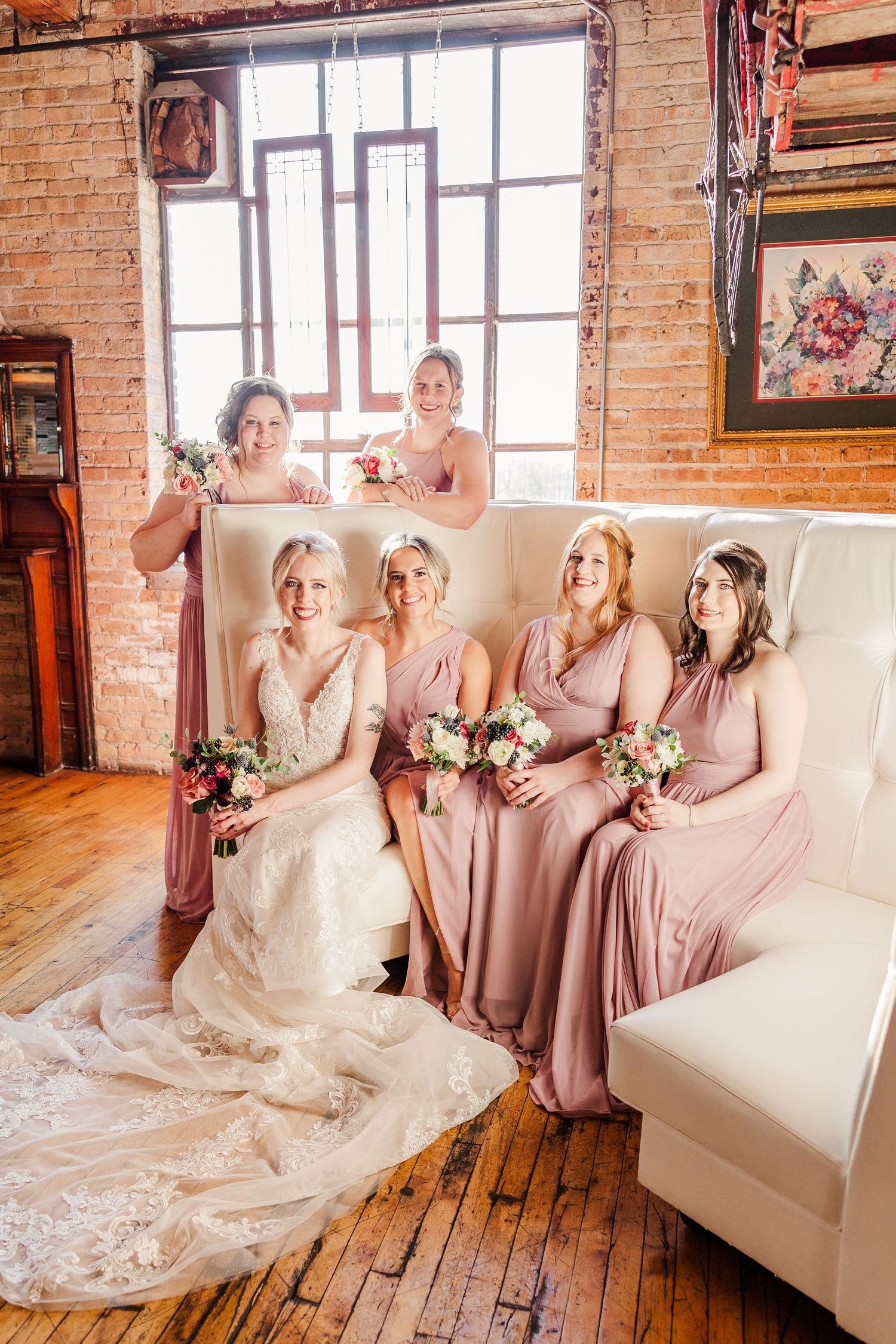Bridal party in rose-colored dresses gathered on white couch in industrial-style room.