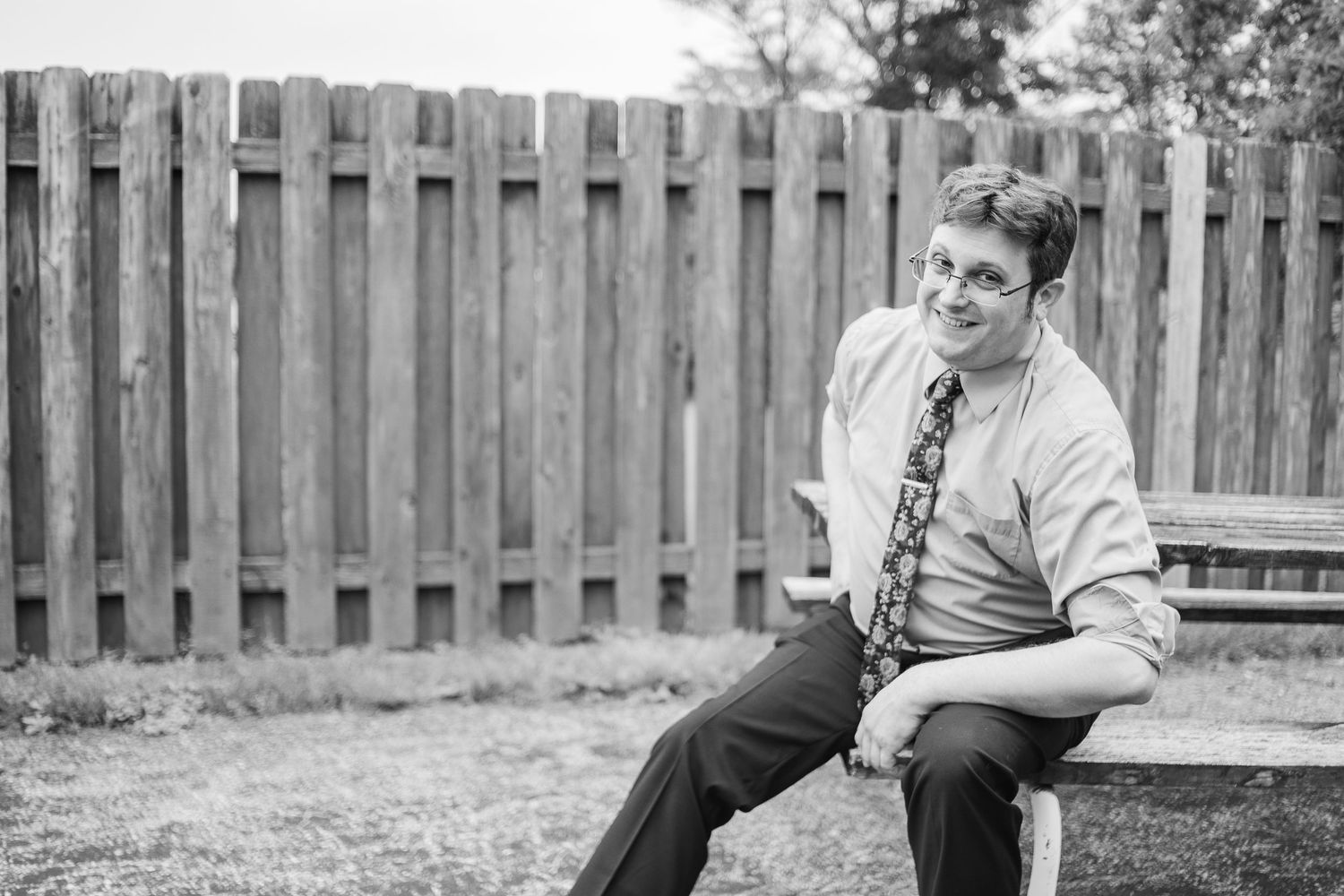 Black and white portrait of someone seated outdoors by a wooden fence.
