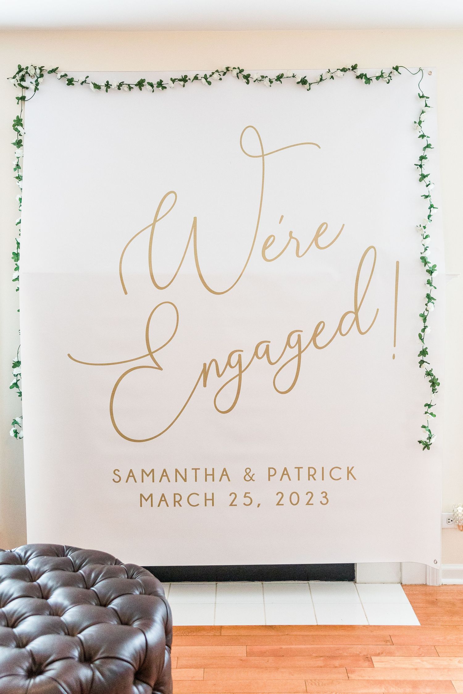 White engagement announcement backdrop with gold script lettering and delicate green vine border.